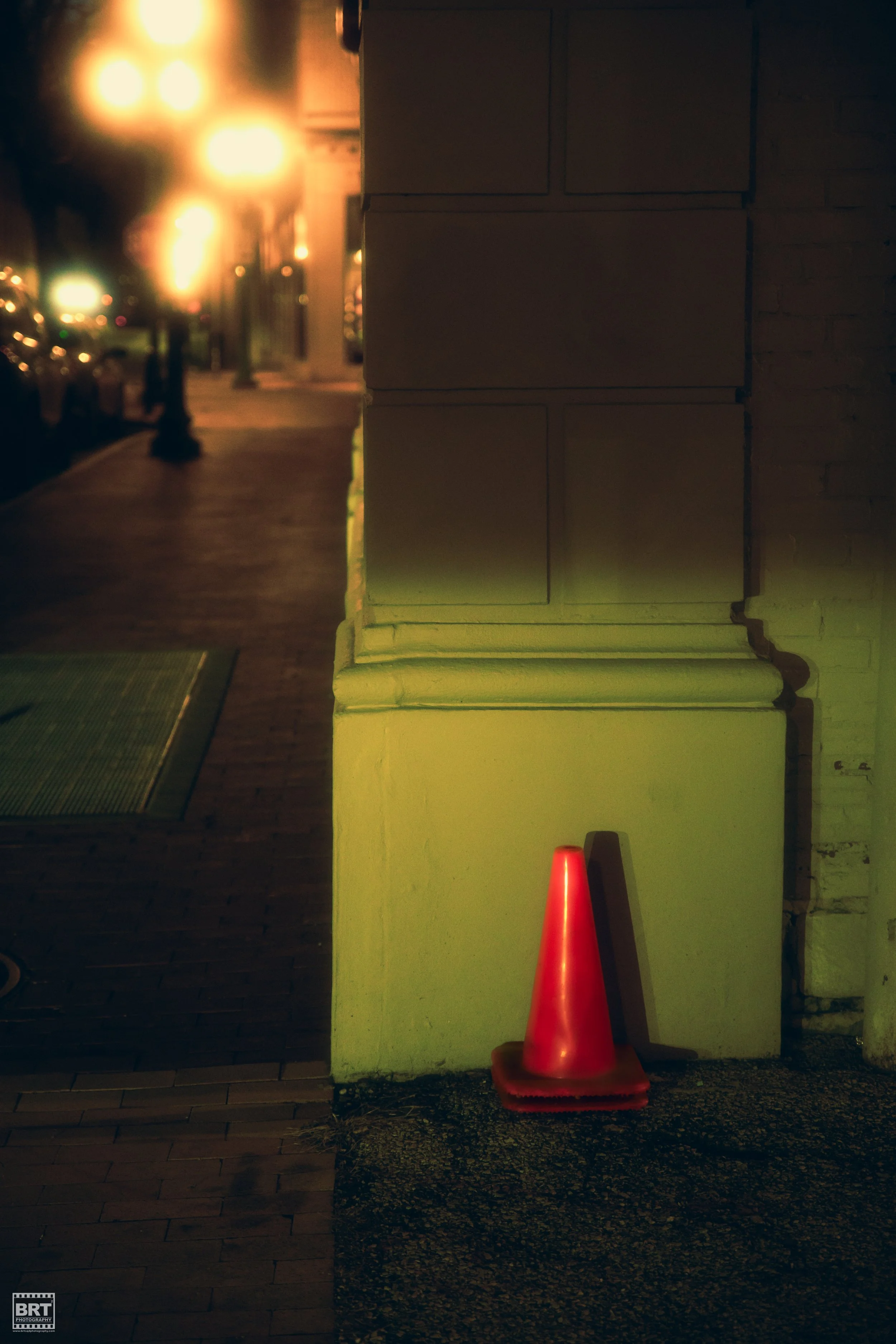 A red traffic cone leaning against a corner of a building with a brick wall and illuminated by streetlights at night.