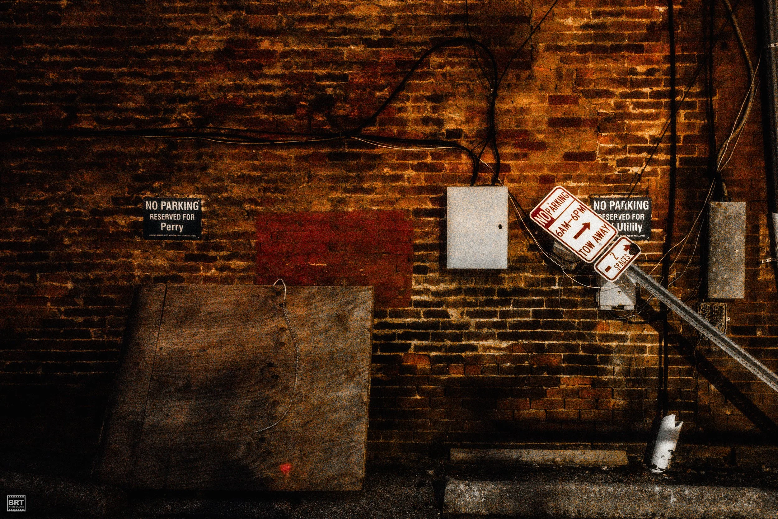 A brick wall with two signs that read 'No Parking Reserved for Perry' and 'No Parking Reserved for Utility'. There is a leaning damaged parking sign, electrical boxes, wires, and a large wooden board leaning against the wall.