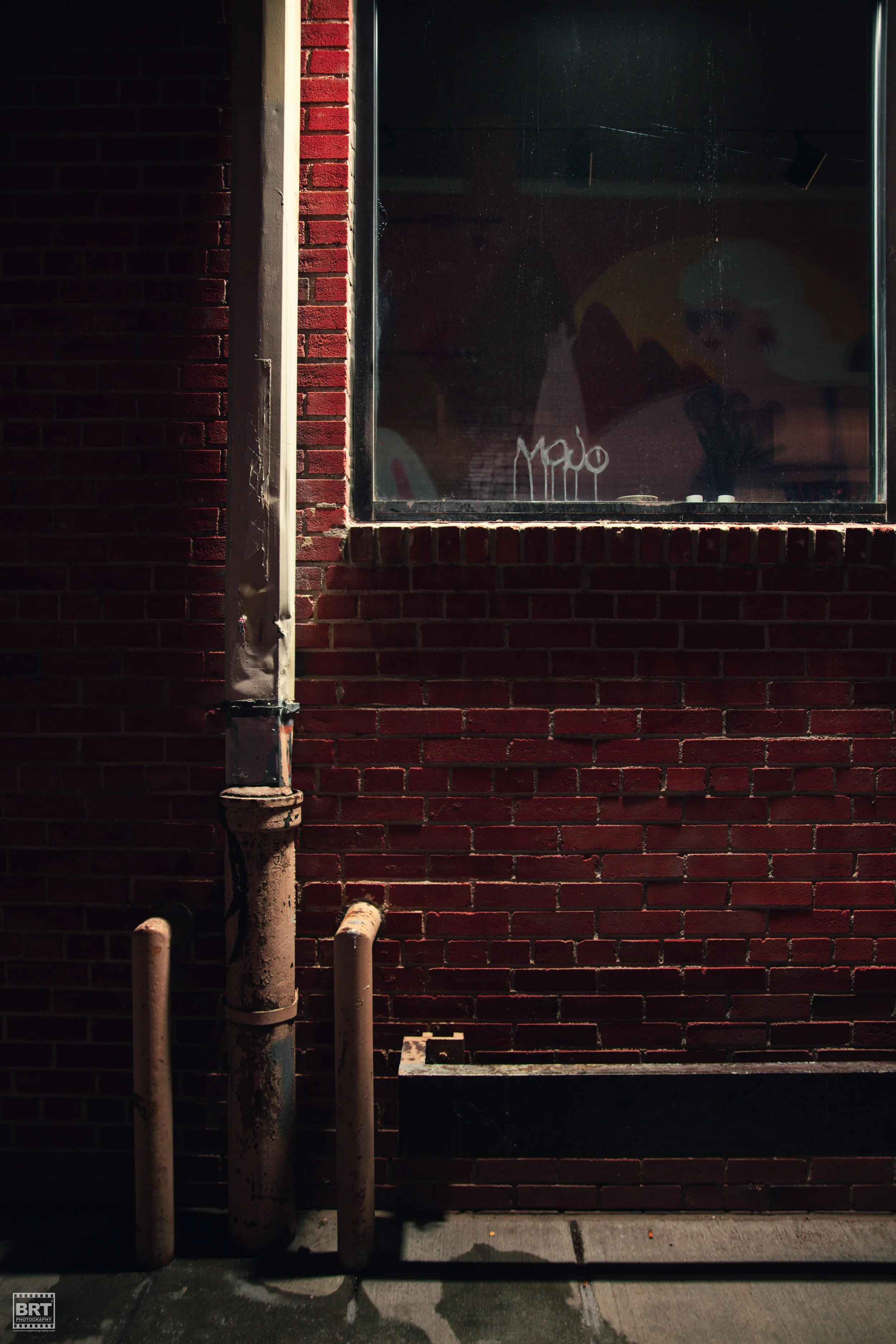 Close-up of a red brick wall with a large window, graffiti reading 'MALO' on the glass, and rusted metal pipes on the sidewalk.