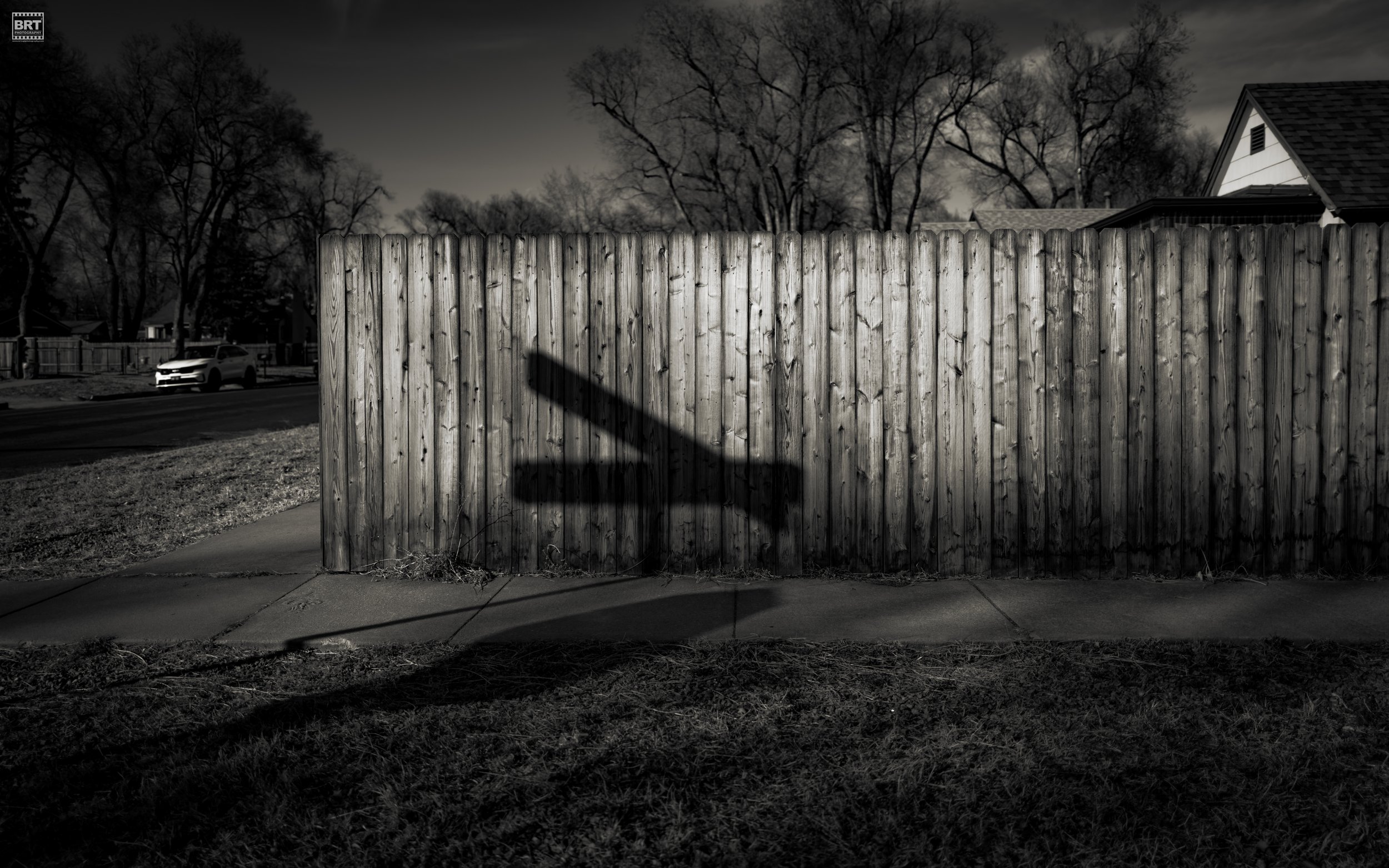 A wooden fence with a shadow of a street sign cast on it, in a neighborhood with houses and leafless trees in the background, under a cloudy sky.