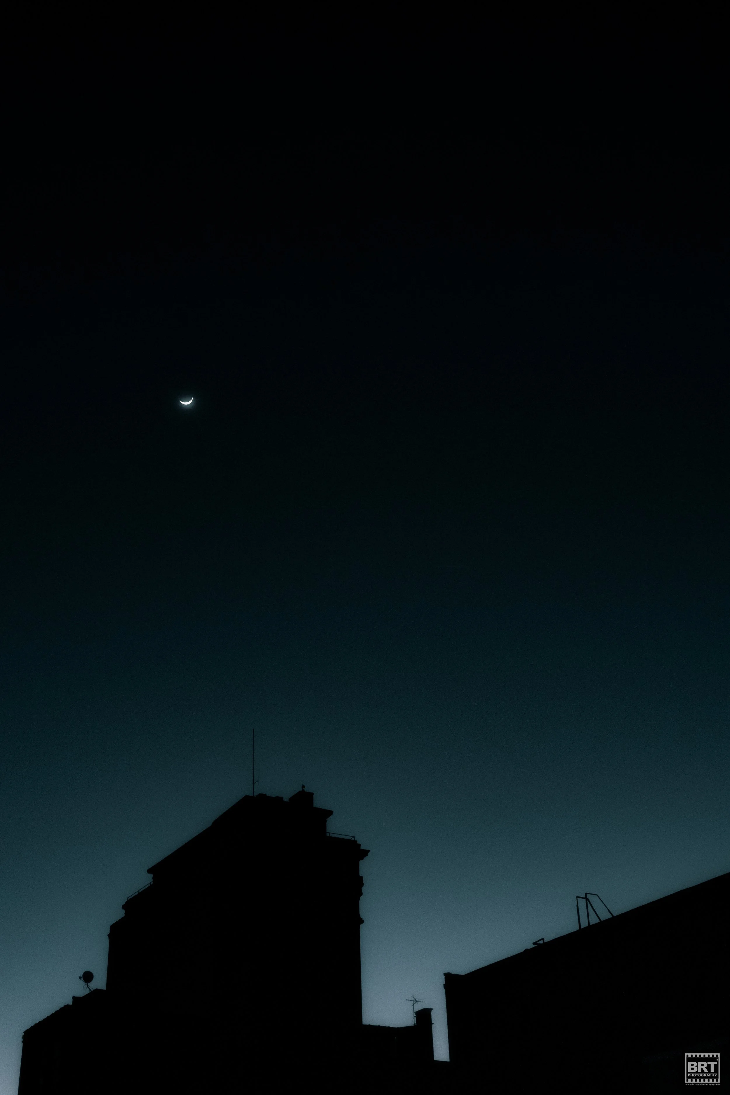 Nighttime cityscape with silhouetted buildings and a crescent moon in the clear sky.