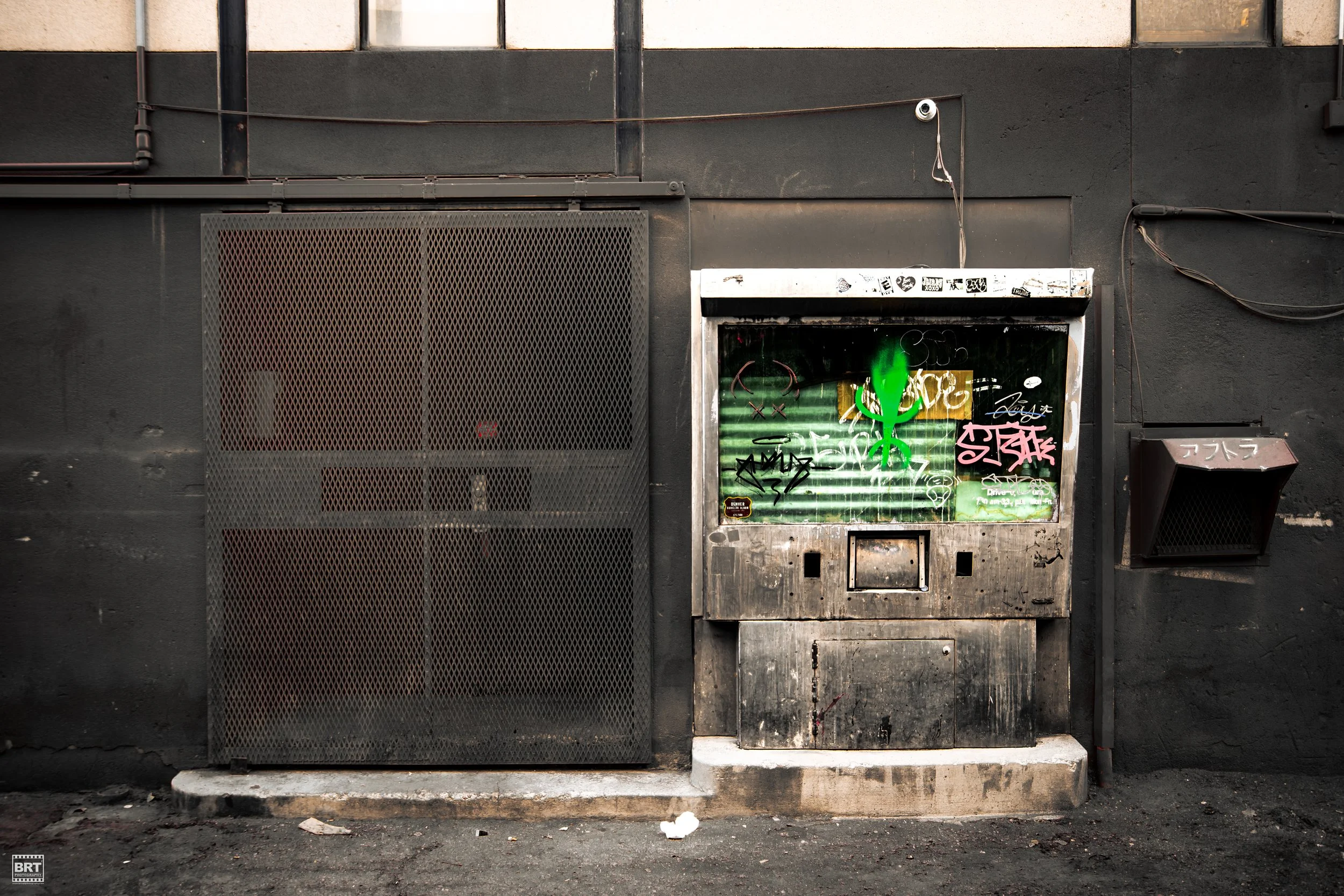 Street scene with a dark wall, metal vent, and graffiti-covered vending machine with a bright green lizard illustration, and graffiti tags in various colors.
