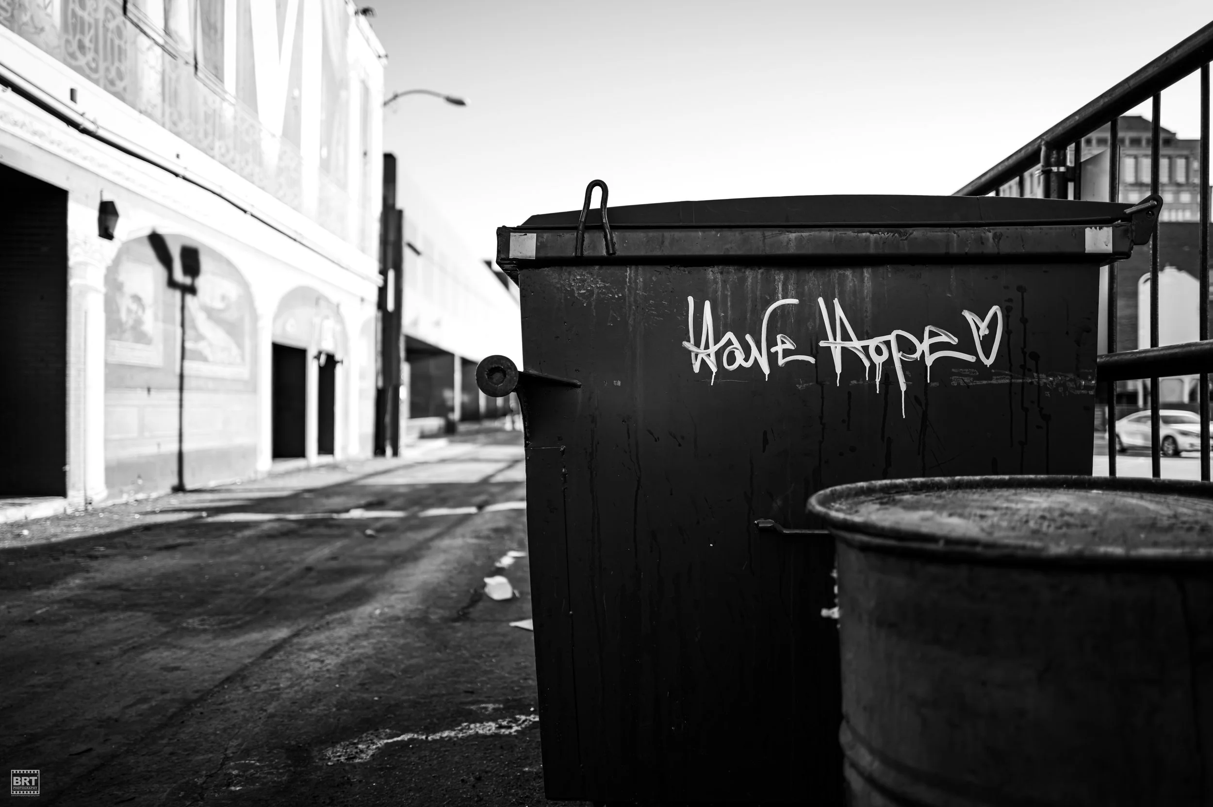 A black garbage bin with graffiti that says 'Have Hope' on an urban street in black and white.