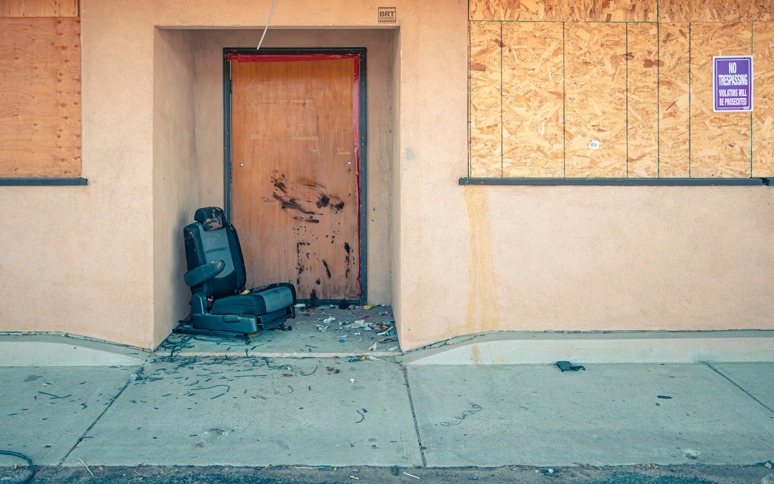 A damaged office chair next to a boarded-up door with black smudges and debris on the ground; plywood covers the window on a beige wall with a purple no trespassing sign.