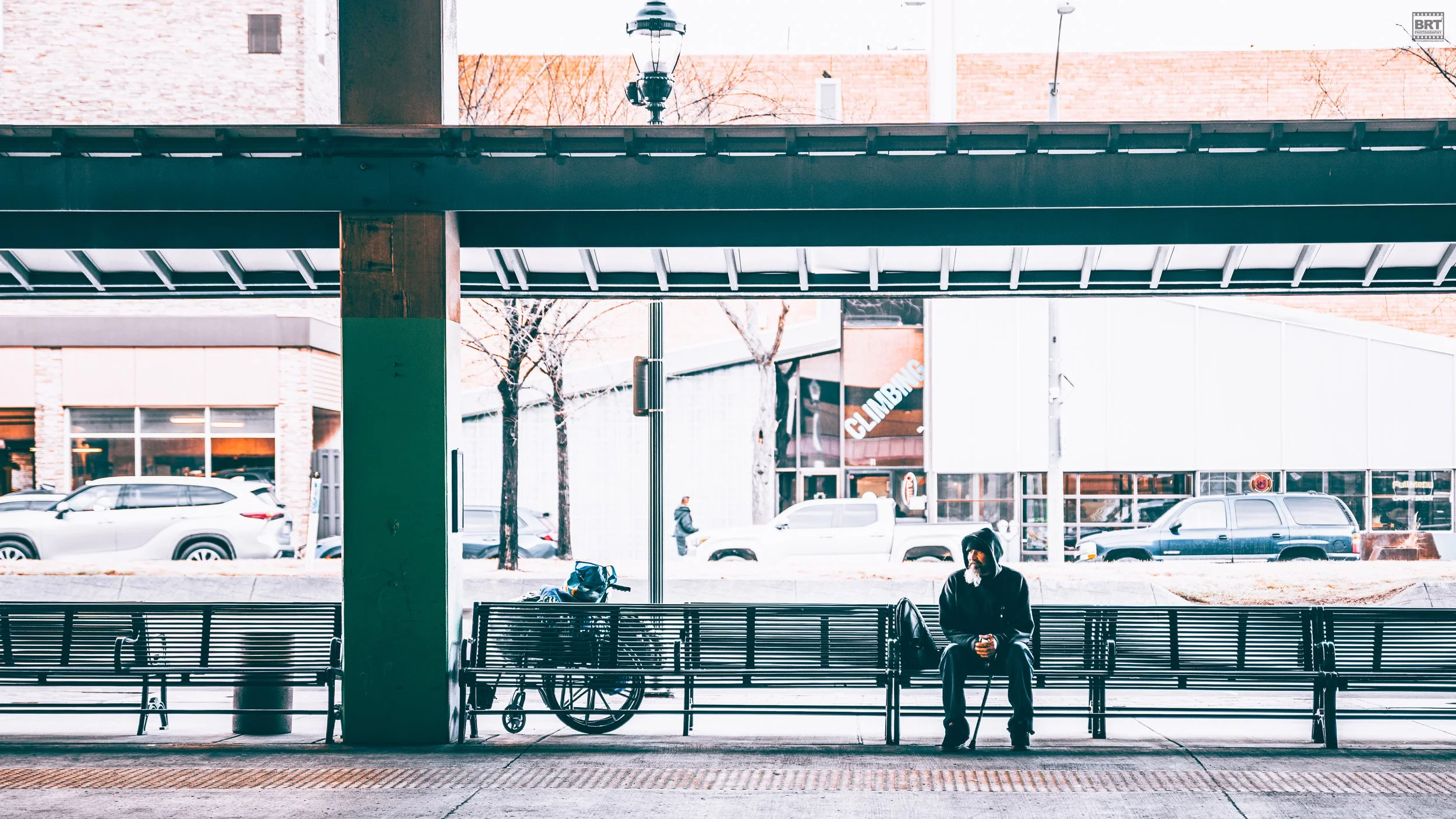 A man with a beard wearing a hoodie and glasses sitting on a bench at a bus stop, with a bicycle and backpack nearby. There are parked cars and a building with a climbing gym sign in the background. The scene is outdoors during winter.