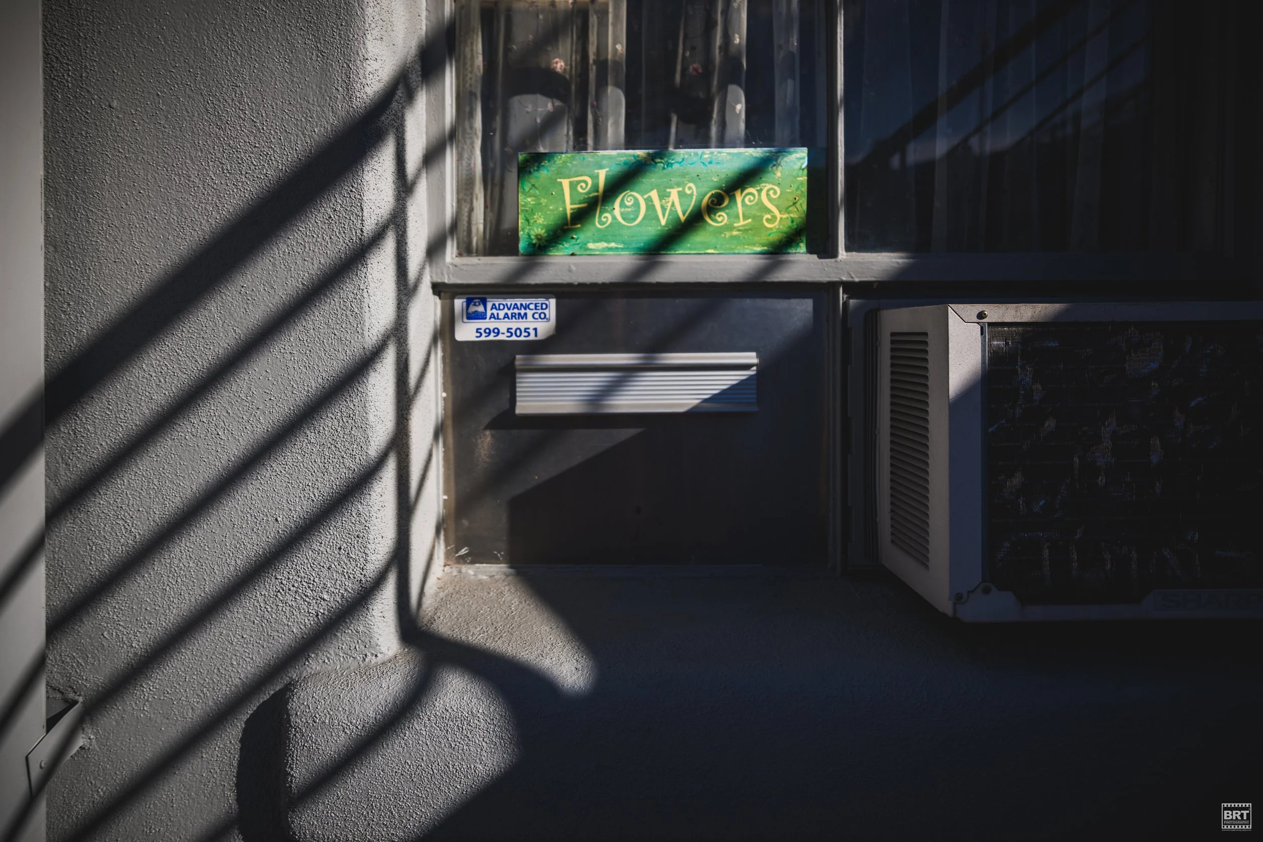 A window with a green sign reading 'Flowers', a blue security sticker, and an air conditioning unit in a partly shadowed outdoor scene.
