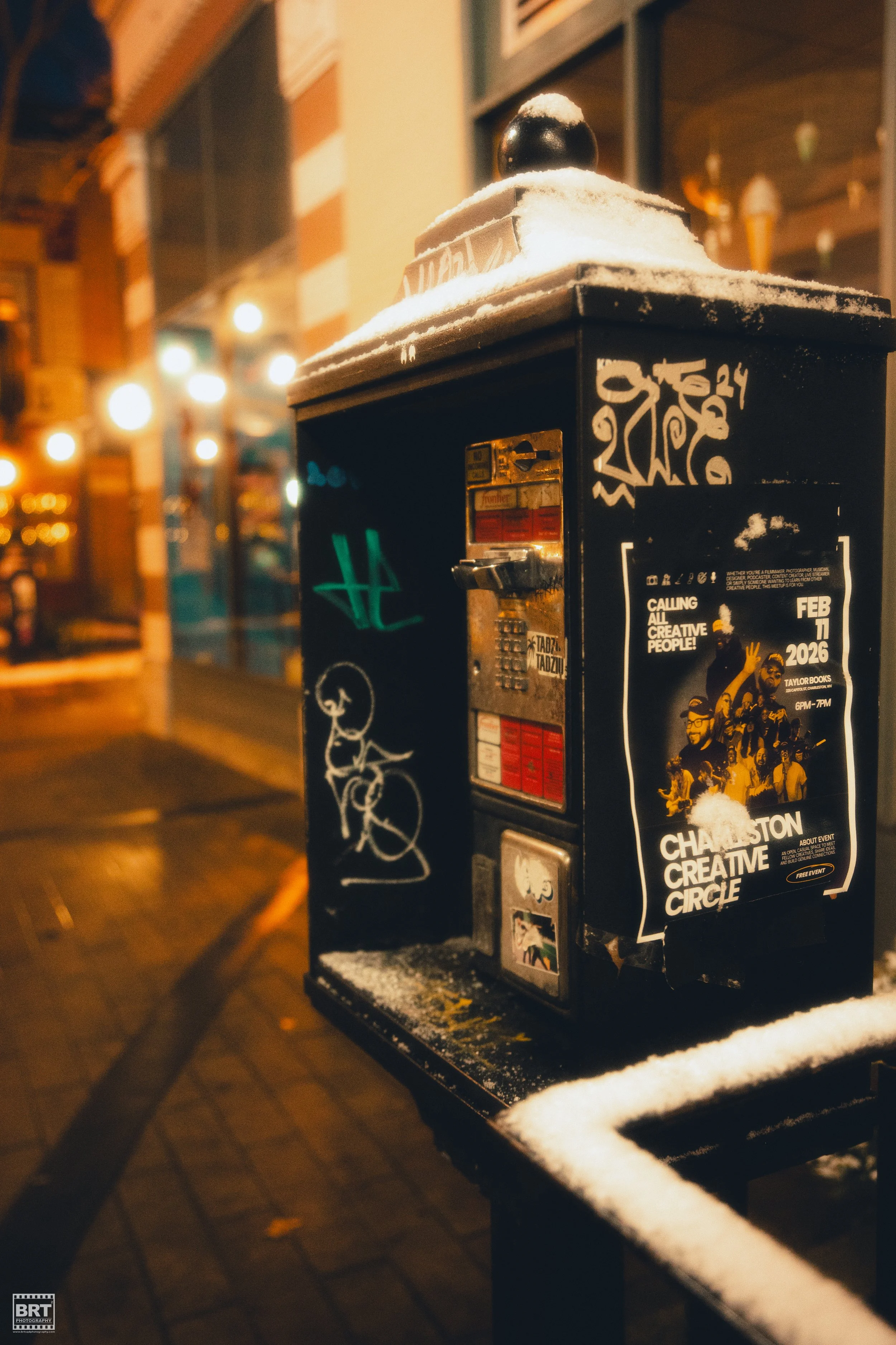 A black payphone booth with graffiti, stickers, and a poster, covered with snow, on a city sidewalk at night with blurred streetlights and buildings in the background.