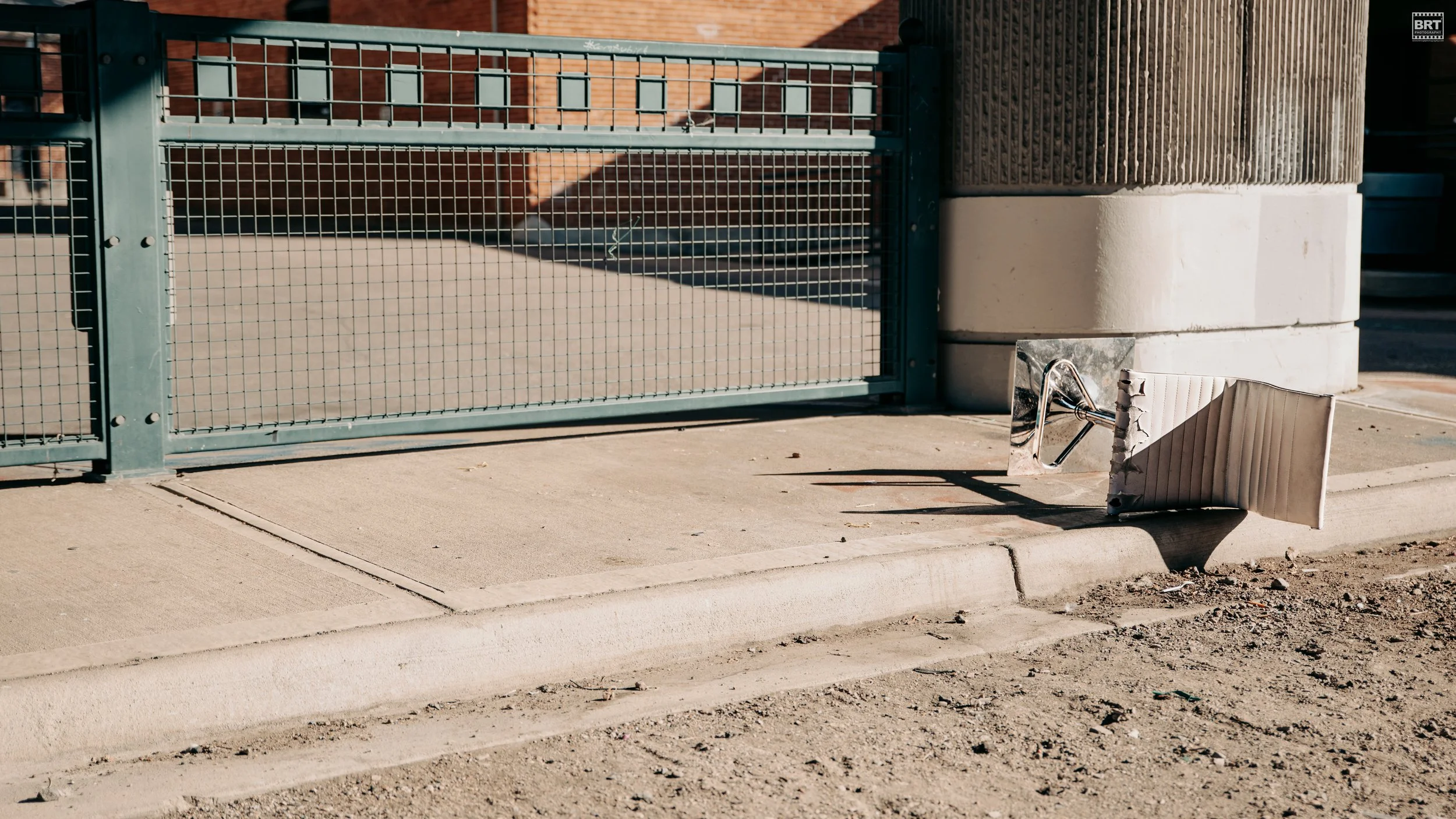 A toppled metal and plastic sign or fixture lying on the sidewalk near a brick building and a metal gate, with dirt and debris on the ground.