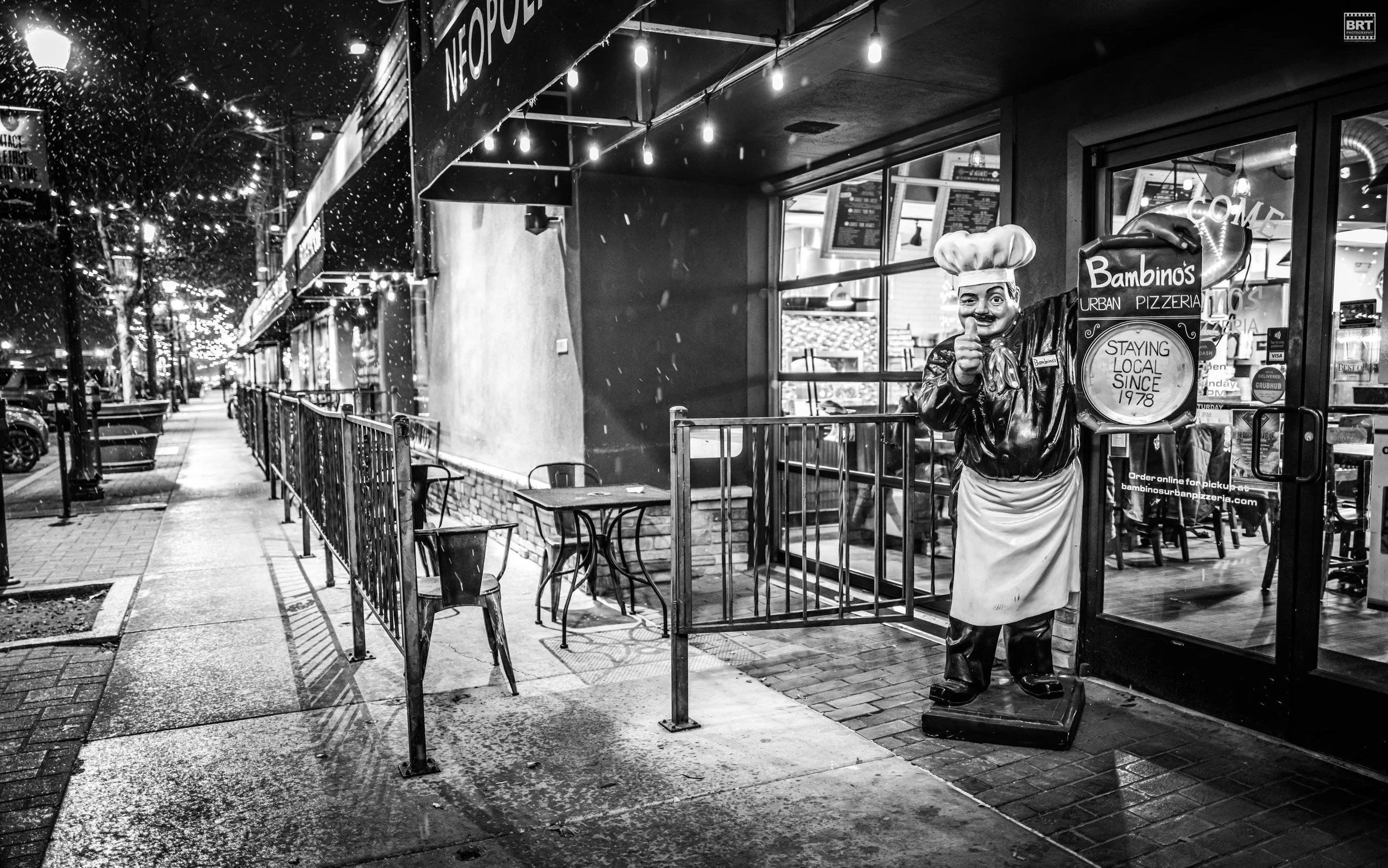 Nighttime view of a sidewalk outside Bambino's Urban Pizzeria, featuring a large chef statue pointing forward near the entrance. The shop has string lights, and empty outdoor tables and chairs are visible.