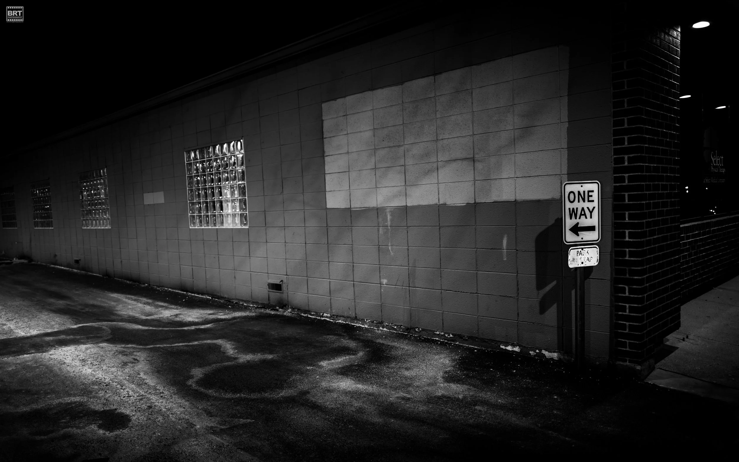 A black and white photo of a building with glass block windows and a parking lot. A sign indicates one-way traffic with an arrow pointing left.