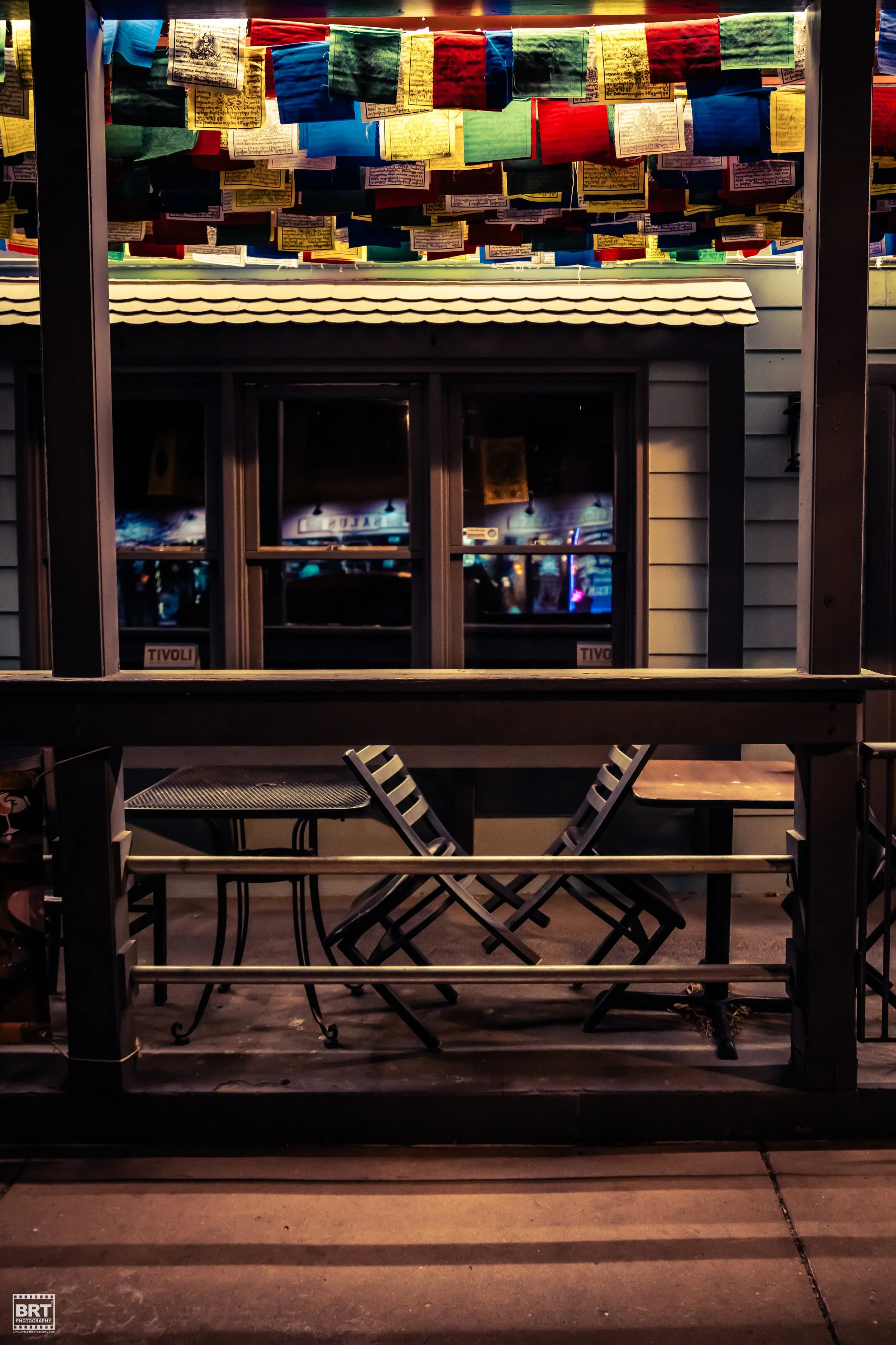 An outdoor seating area with two black chairs and two small tables in front of a house with gray siding. Above, colorful prayer flags are hanging, illuminated by warm lighting, and reflections of a neon-lit street scene are visible through the window