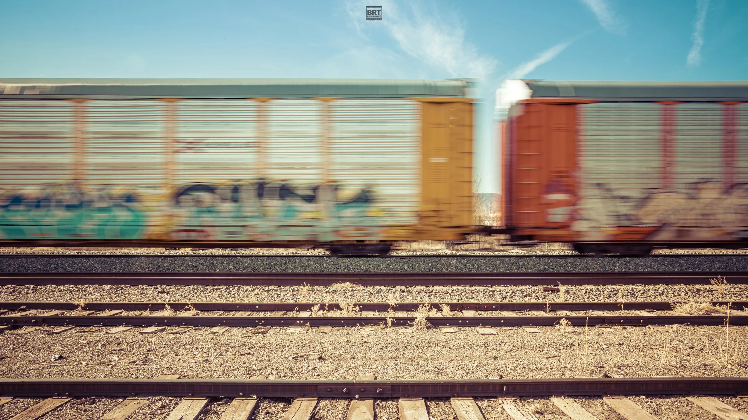Blurred freight train cars moving on railway tracks under a clear blue sky with some clouds.