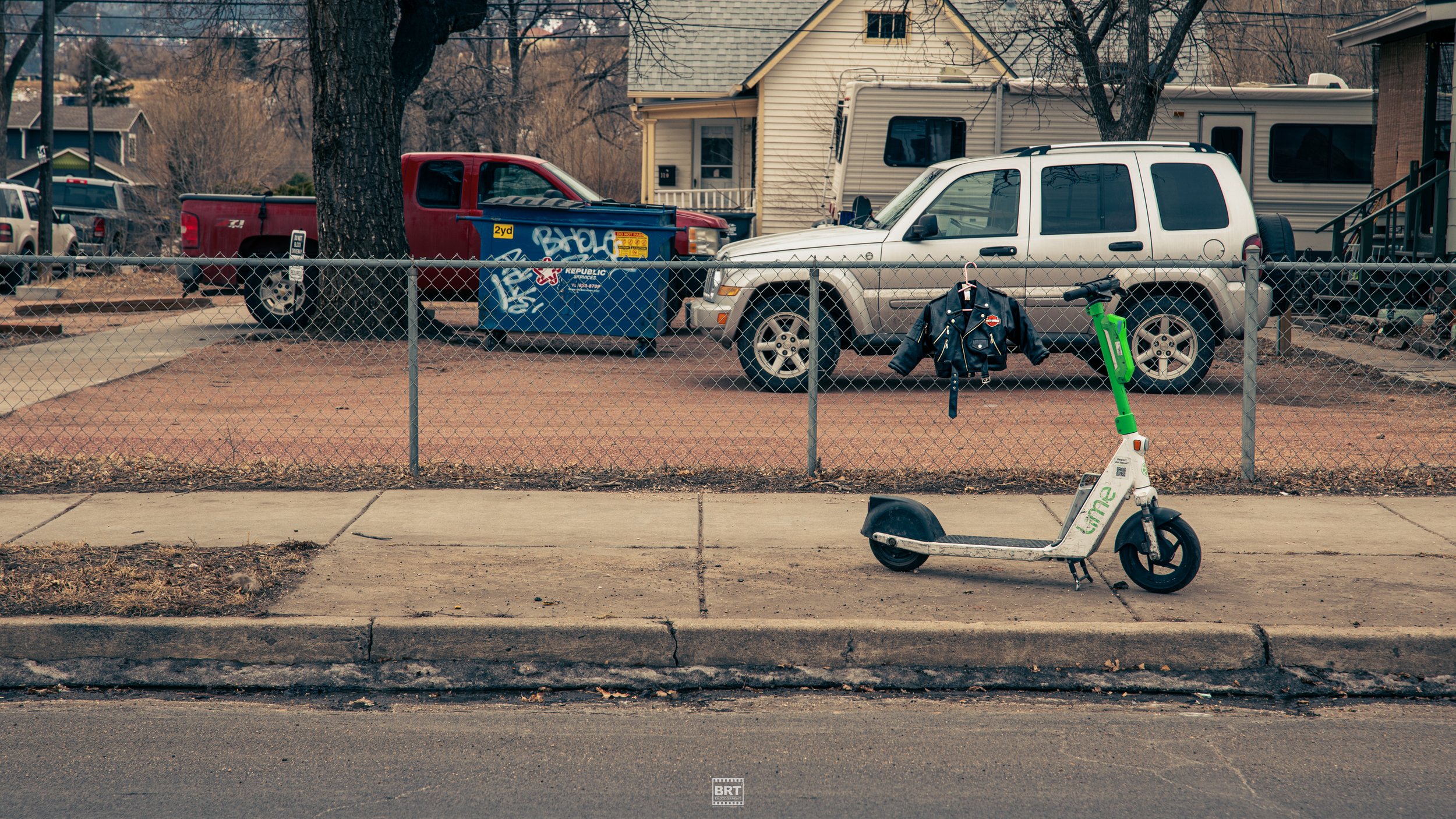 An electric scooter parked on a sidewalk in front of a chain-link fence. Behind the fence, there is a house with a yard, parked vehicles including a white SUV, a red pickup truck, and a beige trailer. A black jacket is hanging on the fence with a gre