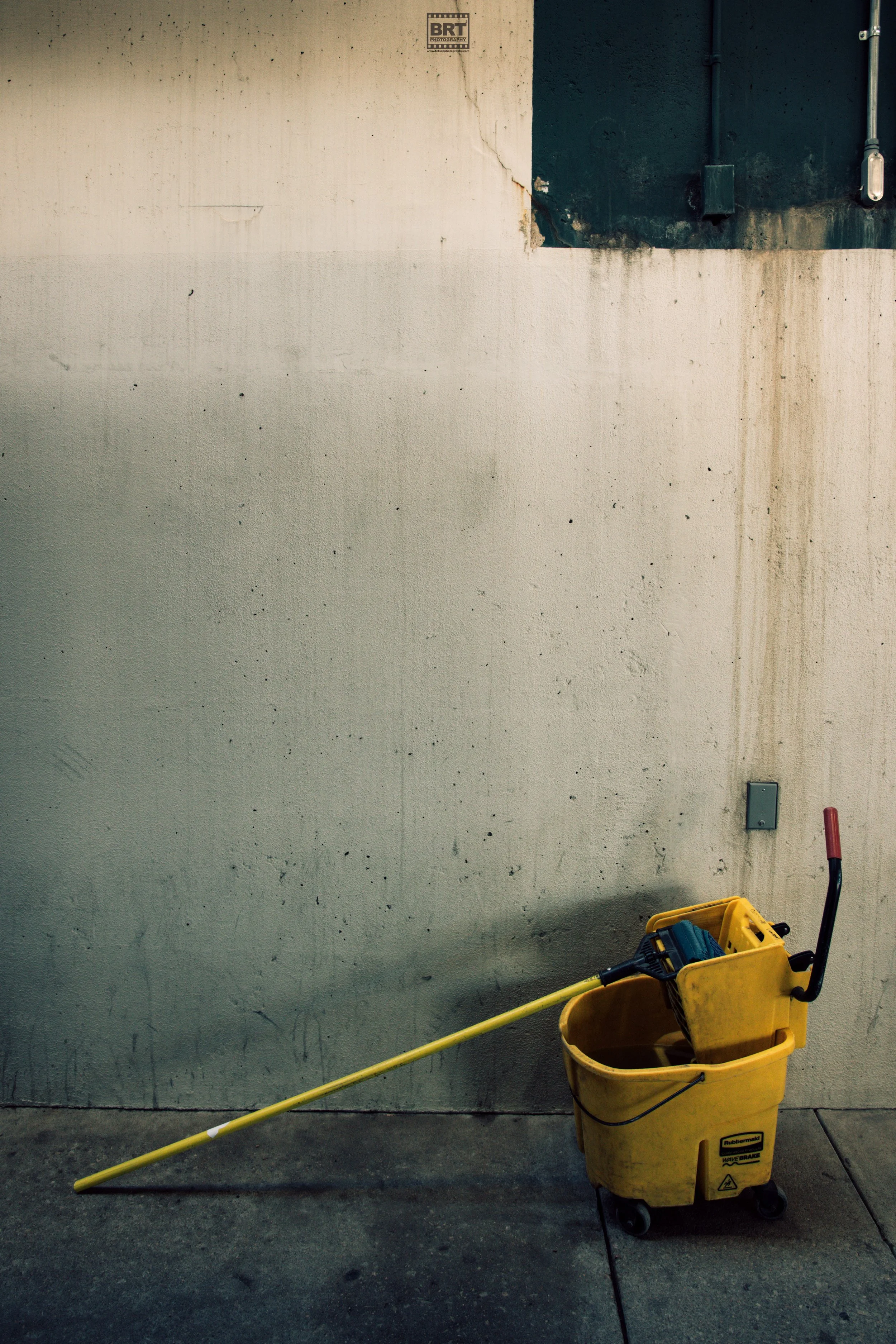 A yellow cleaning bucket with a mop and a squeegee inside, placed against a concrete wall on a dirty floor.