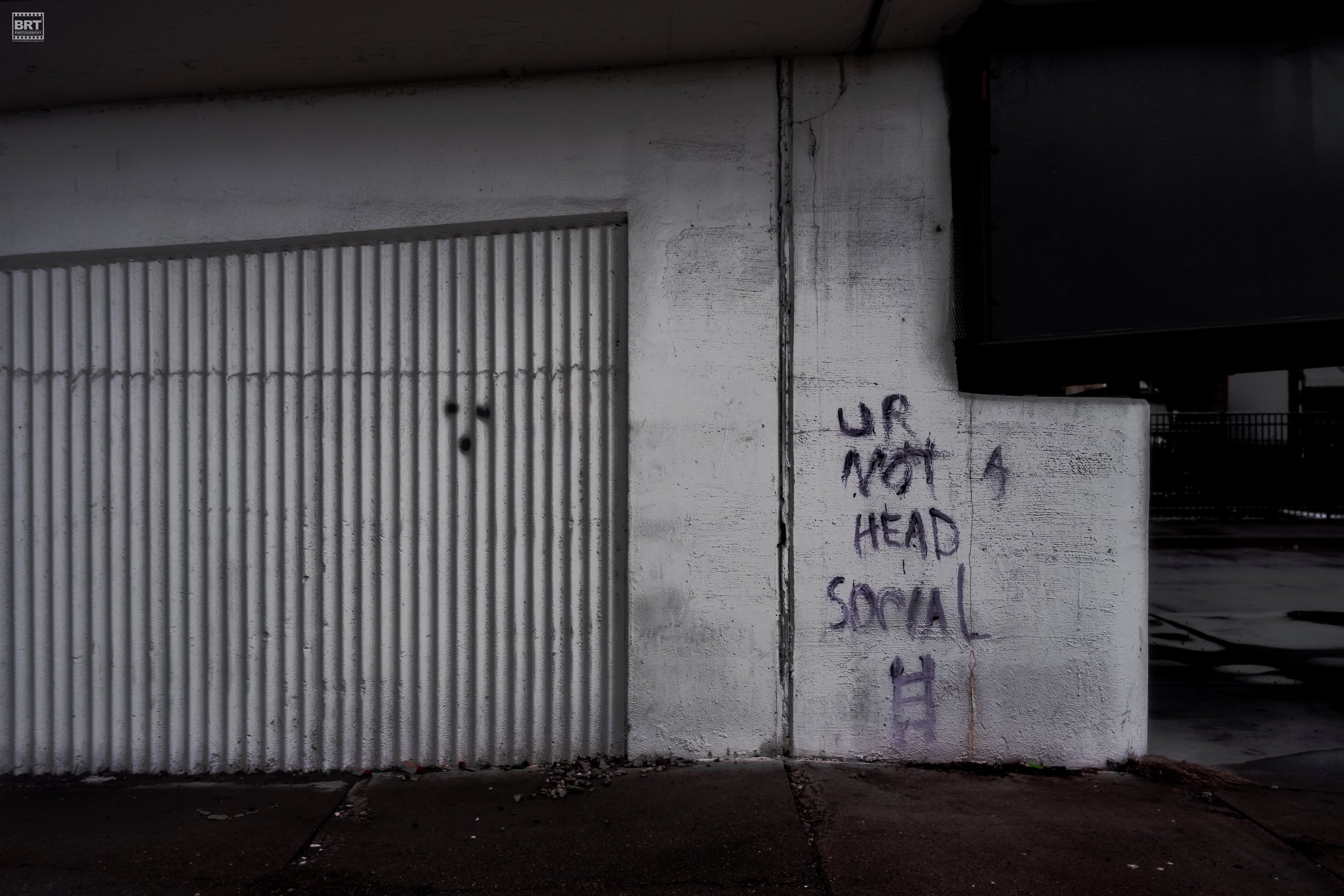Graffiti on a white concrete wall that says 'UP NOT A HEAD SOCIAL' next to a corrugated metal door in a parking garage.