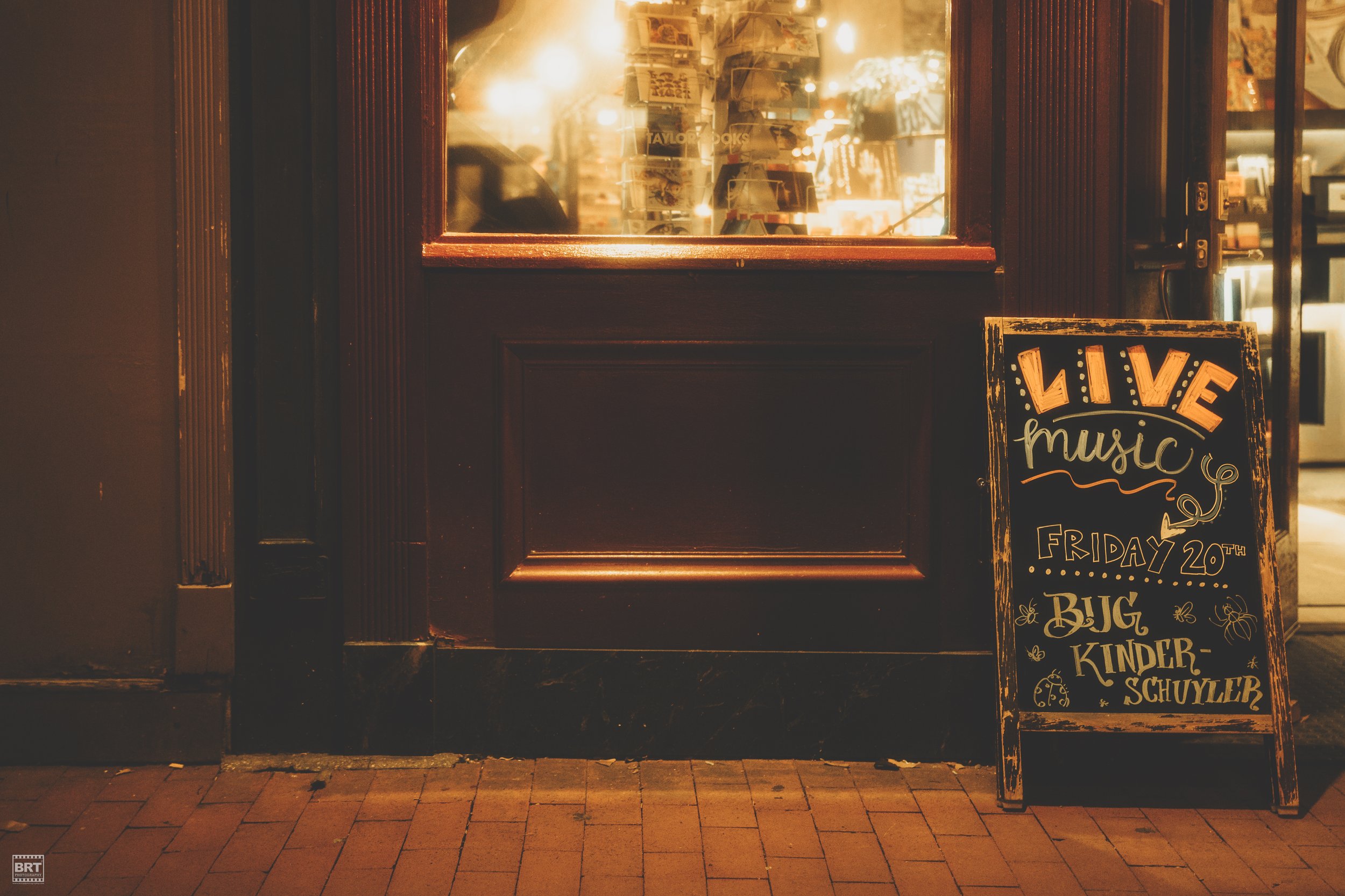 A chalkboard sidewalk sign advertising live music on Friday the 20th at Bug Kinder Schuller, positioned outside a shop window at night. The shop has dark wooden trim, and the interior is warmly lit with items visible inside.