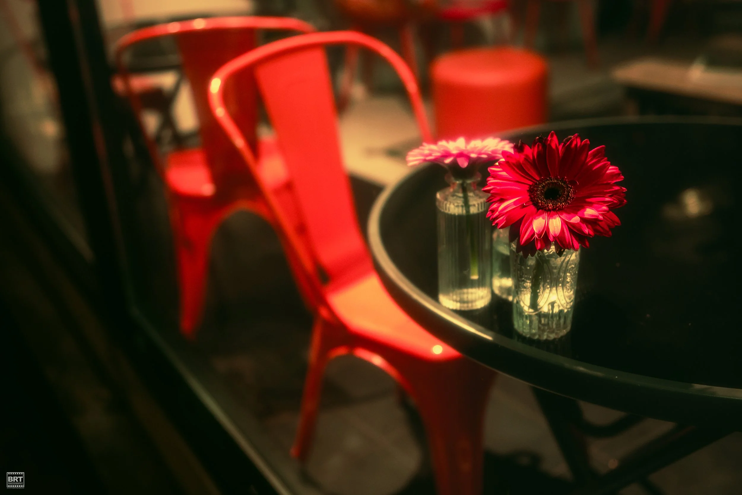 Two vases with flowers on a dark round table, with red chairs in the background