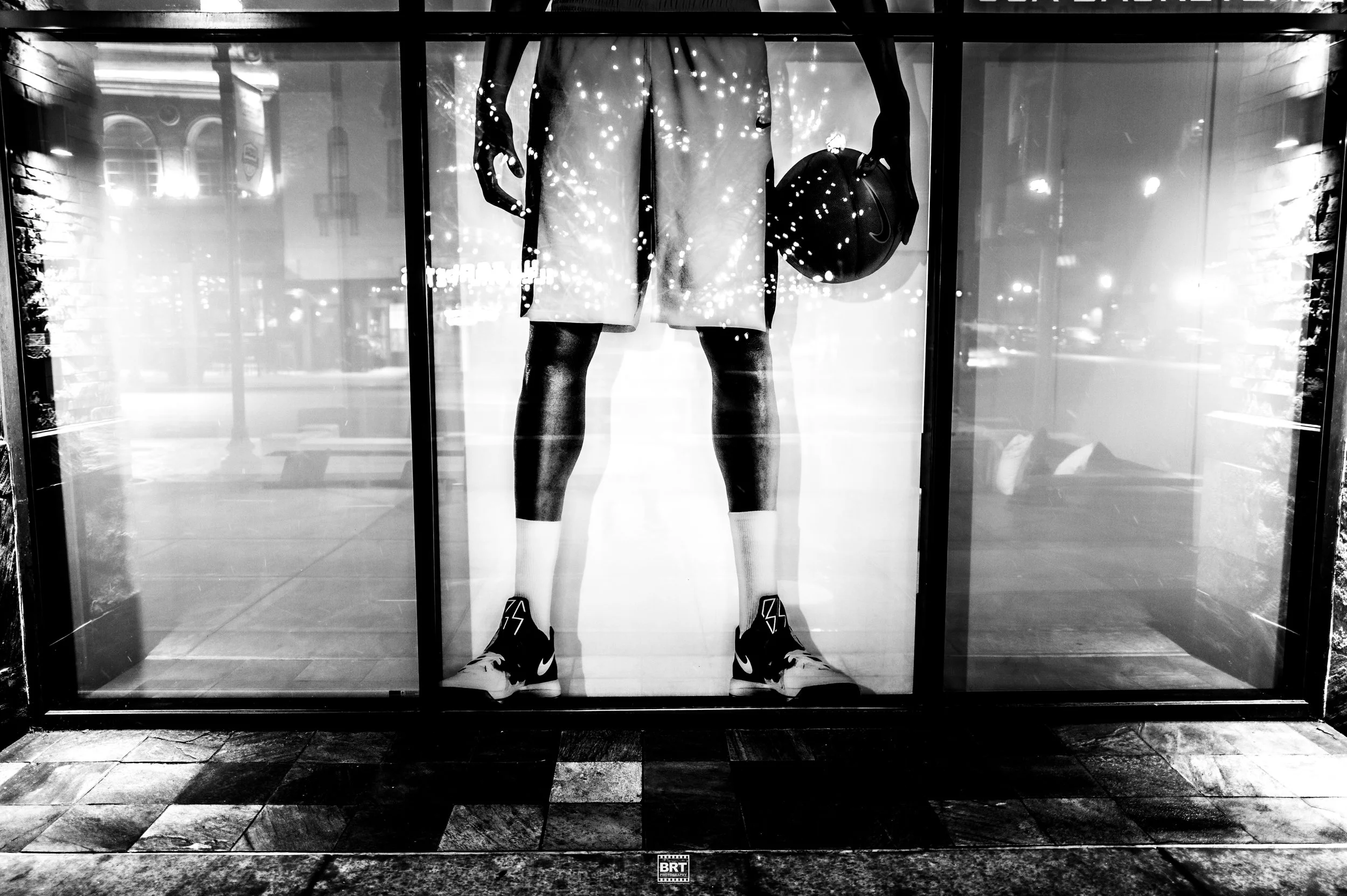 Black and white image of a store window display with a mannequin wearing athletic shorts, shoes, and a t-shirt holding a basketball. The reflection of city lights and buildings can be seen in the window.