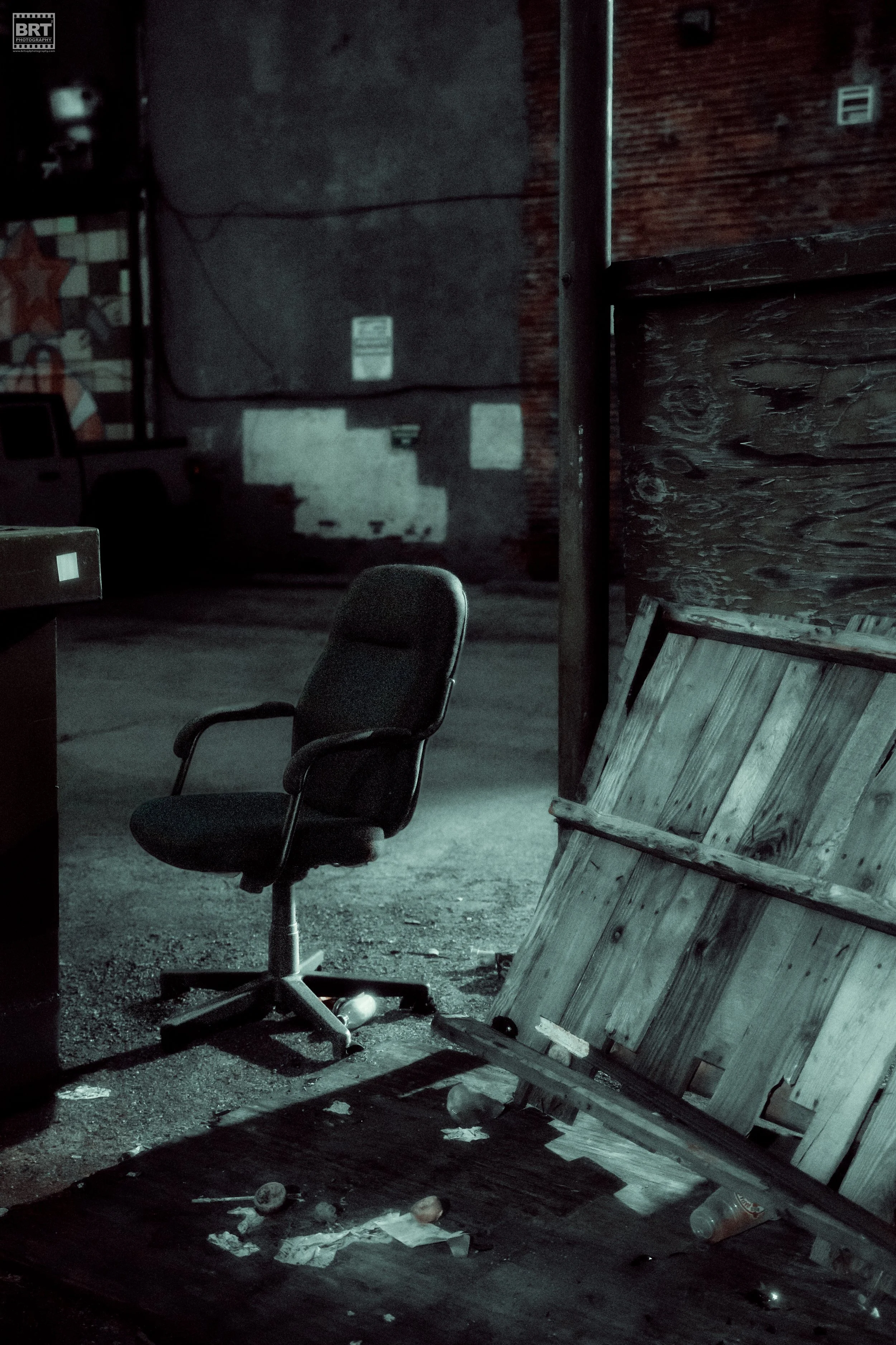 An empty office chair, a wooden pallet leaning against a pole, and trash scattered on the ground in a dimly lit outdoor alley at night.