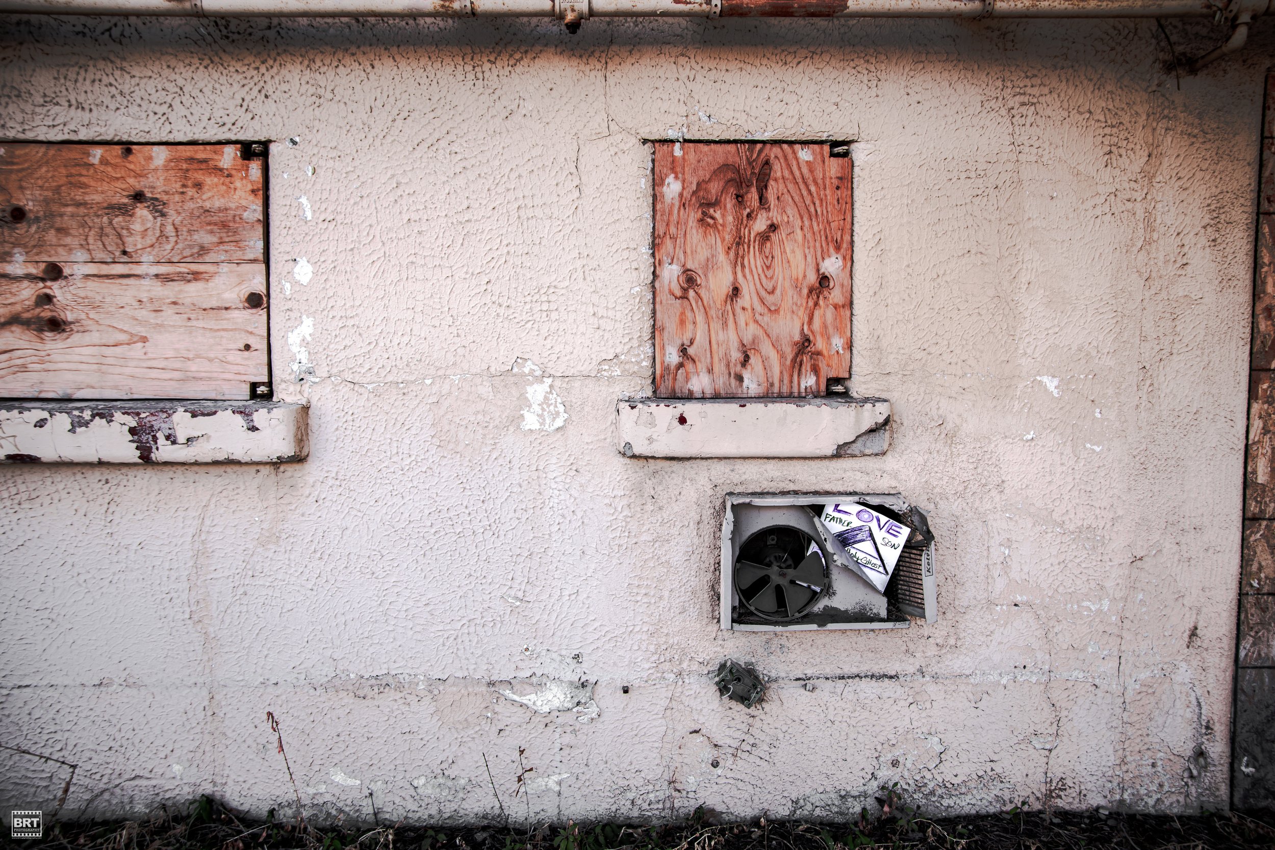 A beige stucco wall with two boarded-up windows covered with plywood and a small broken ventilation opening with a torn flyer inside.
