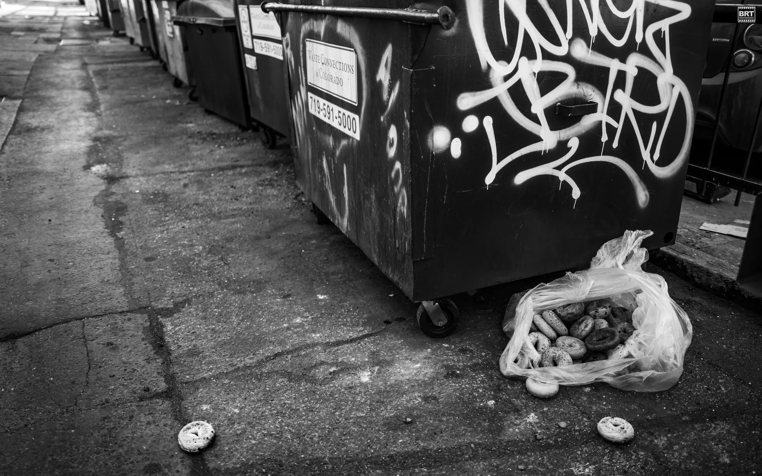 A city street with a row of trash dumpsters covered in graffiti. One of the dumpsters has a plastic bag filled with donuts on the ground next to it, with some donuts scattered on the pavement.