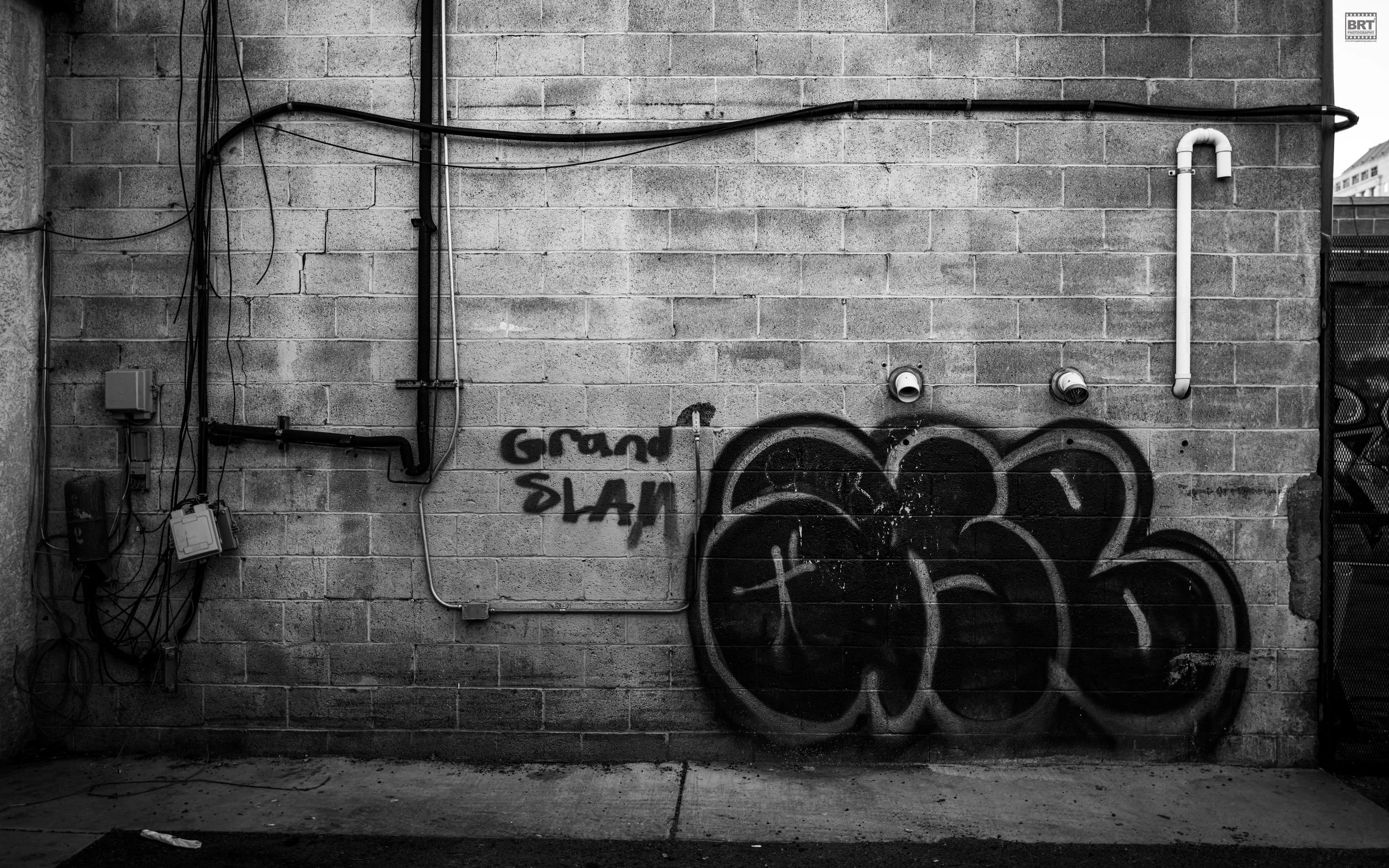 Black and white photo of a brick wall with graffiti reading 'Grand Slam' and large bubble-style graffiti tag, along with electrical conduits and pipes on the wall.