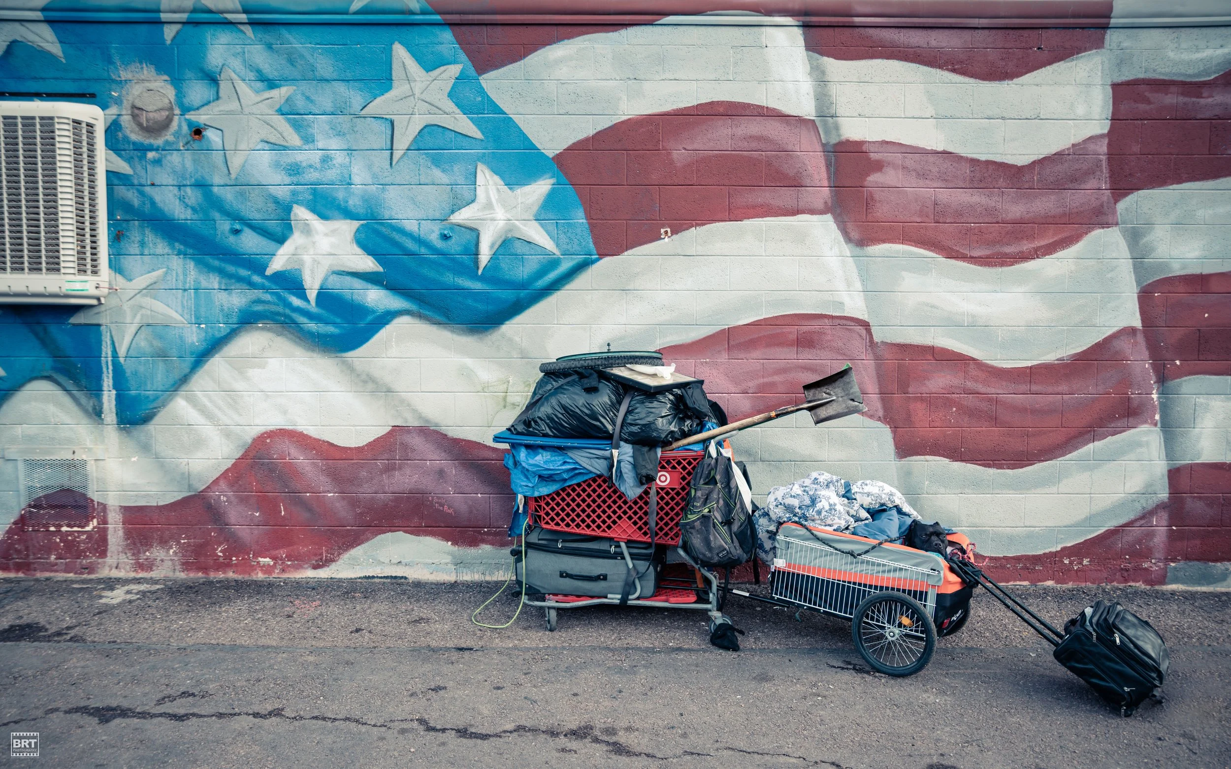 A pile of belongings, including bags, a shopping cart, an umbrella, and a small cart on wheels, is placed against a wall painted with an American flag mural.