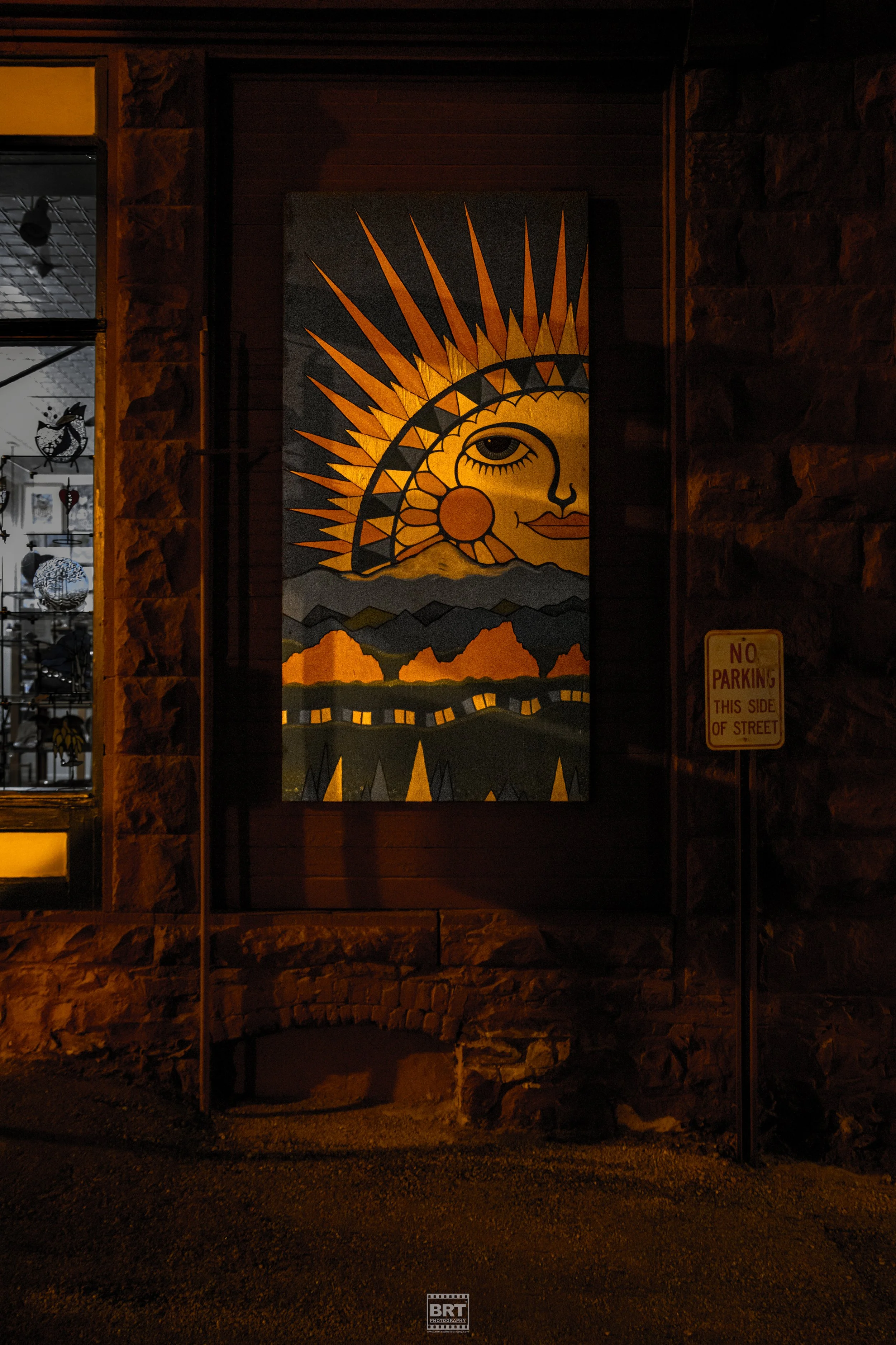 Nighttime photo of a colorful painting featuring a stylized sun with a human face, mountains, and a landscape on a building exterior wall.