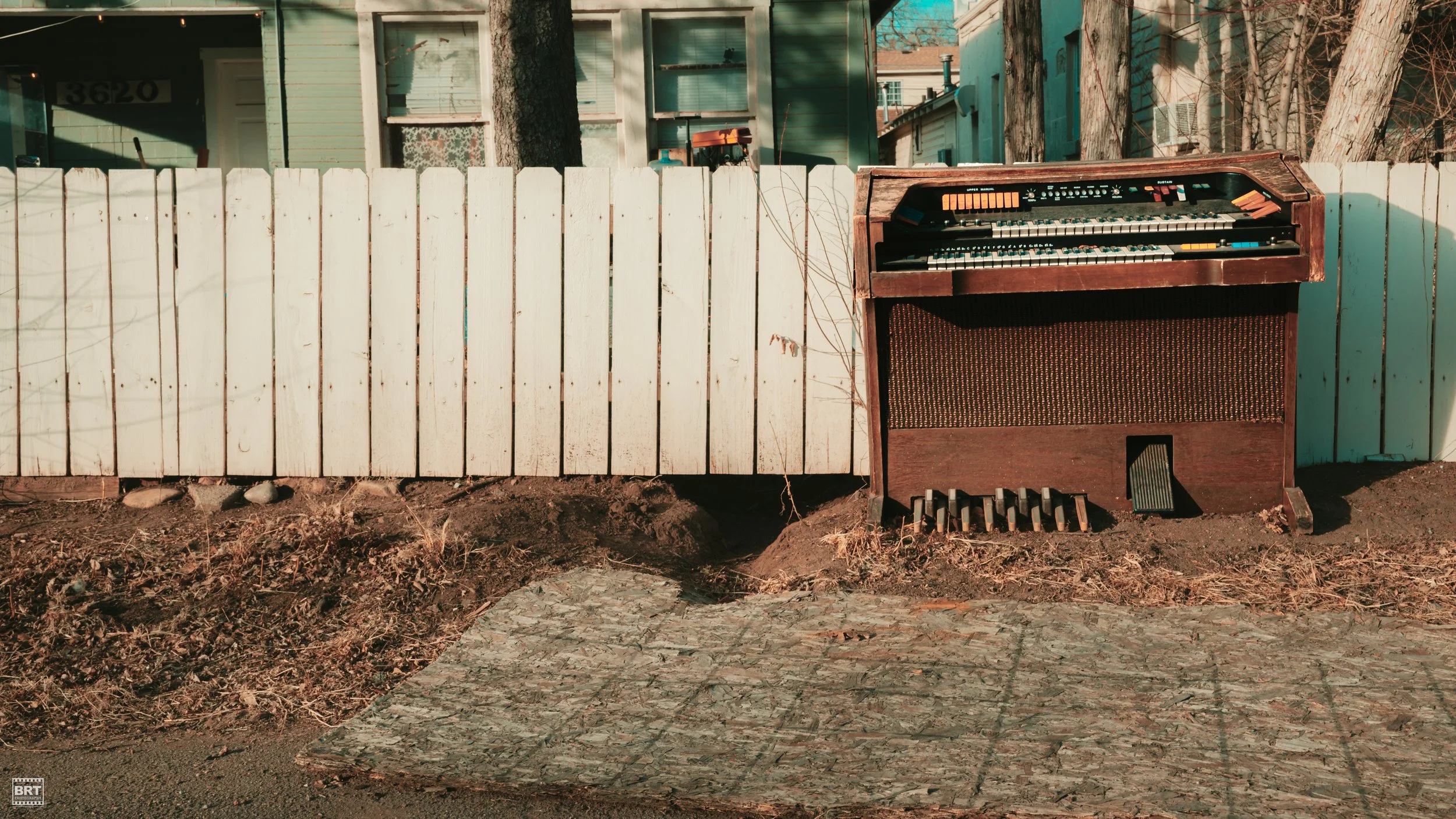 An old electronic organ placed outdoors against a white picket fence, with a partial view of houses and trees in the background.