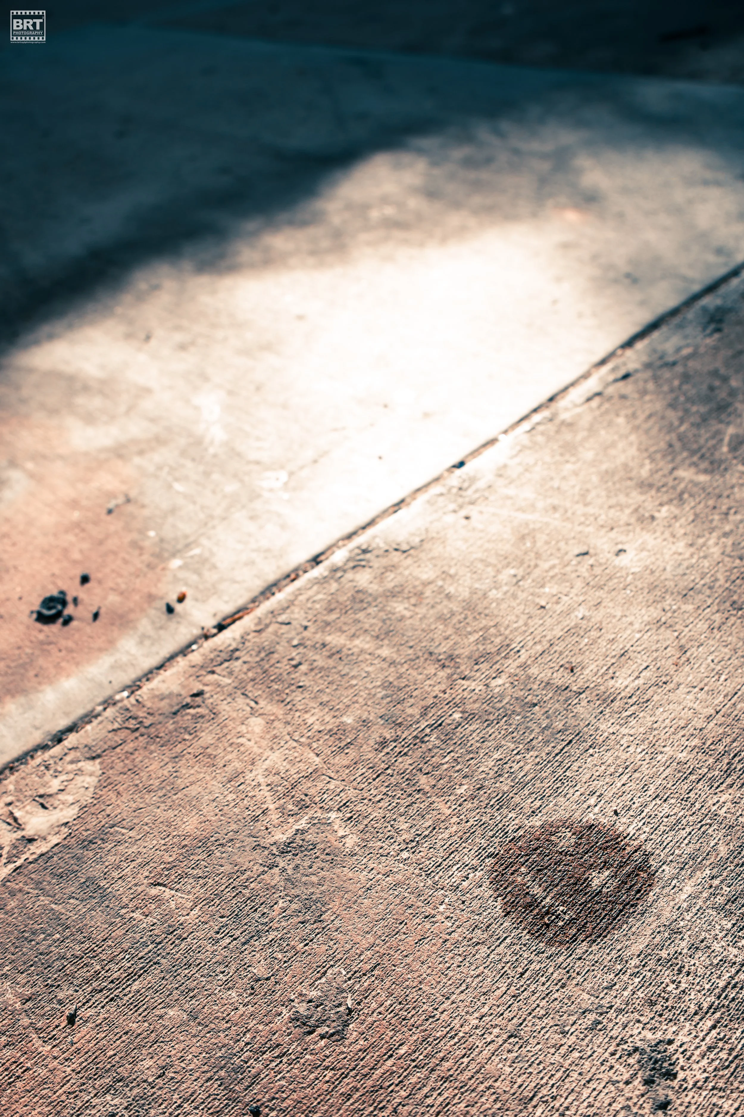 Close-up of a dusty, weathered concrete floor with a dark circular stain near the bottom right, and sunlight casting shadows on the left side.
