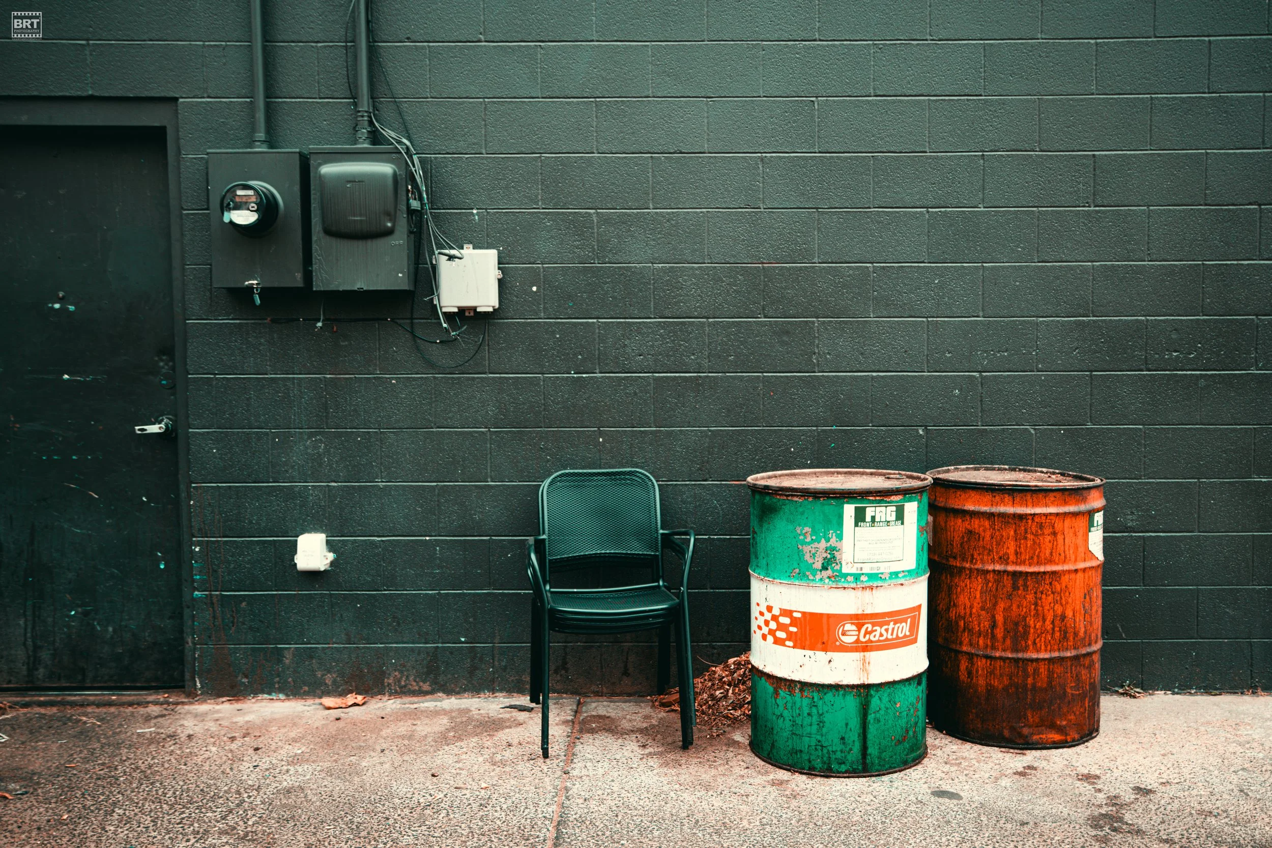 A black brick wall with a black door on the left, and various utility boxes and meters mounted on the wall. In front, there is a single black plastic chair next to two rusty metal barrels, one green and one orange, on a paved ground.