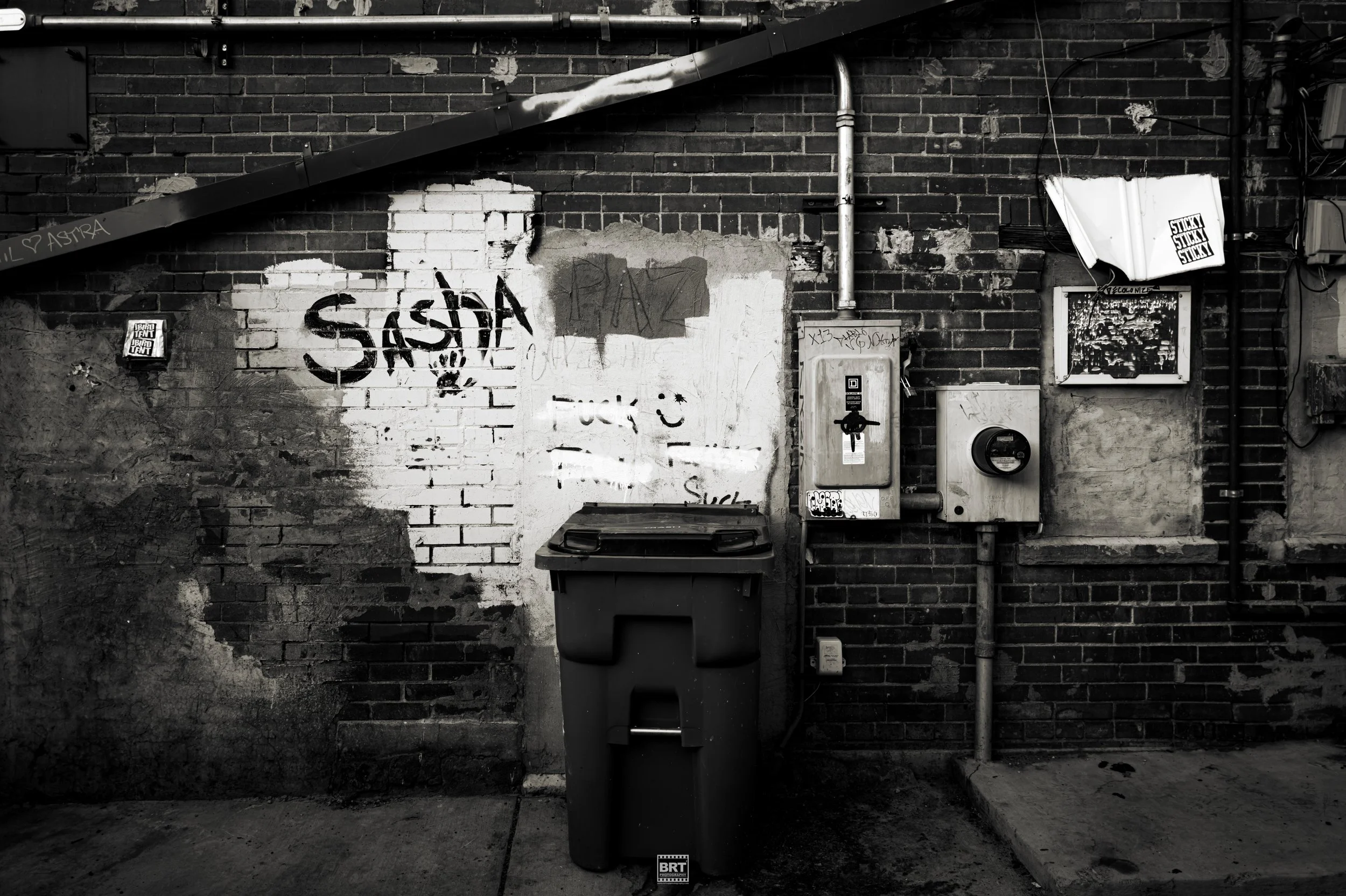 Black and white photo of a brick wall with graffiti that reads 'SASSA,' a trash bin, and electrical boxes. Graffiti and stickers are visible on the wall.