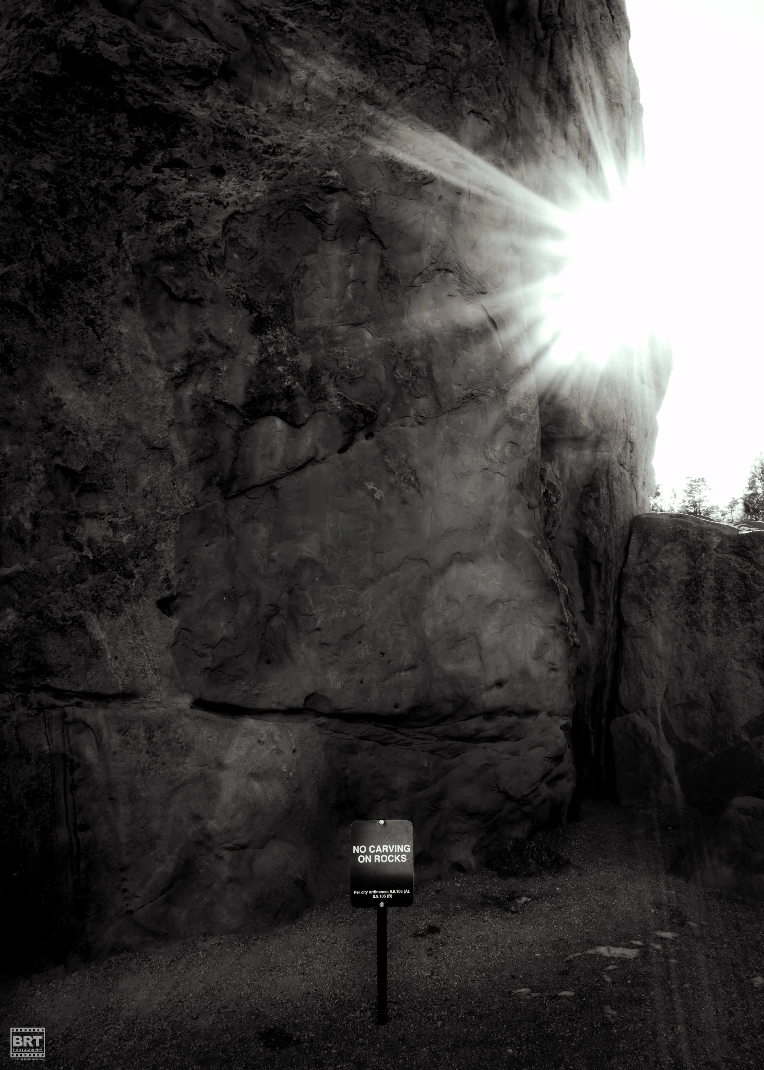 Large natural rock formation with sunlight shining over the top right. A sign at the base of the rocks reads 'NO CARVING ON ROCKS.'