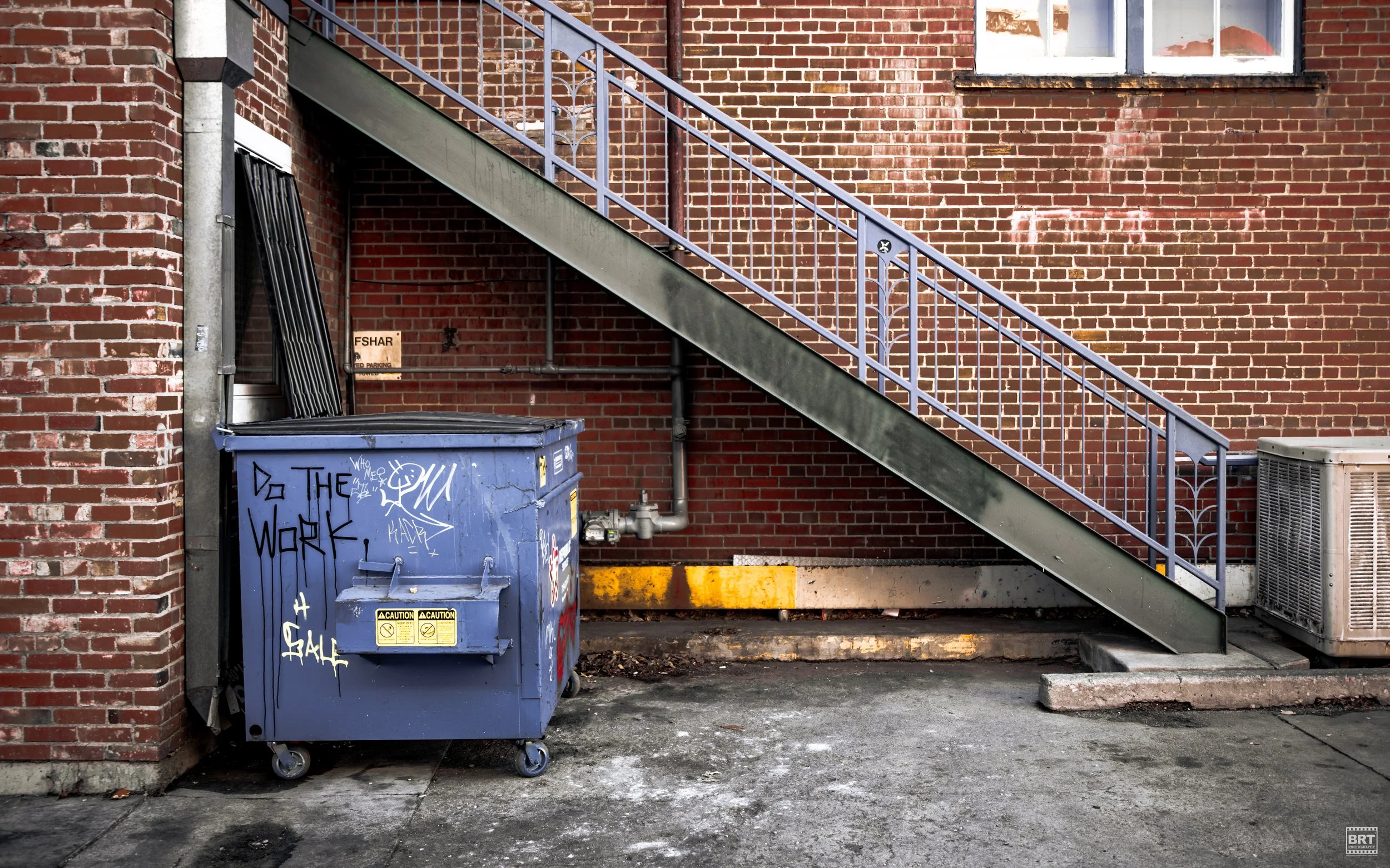 Urban alley with brick building, metal staircase, blue graffiti-covered dumpster, and air conditioning unit.
