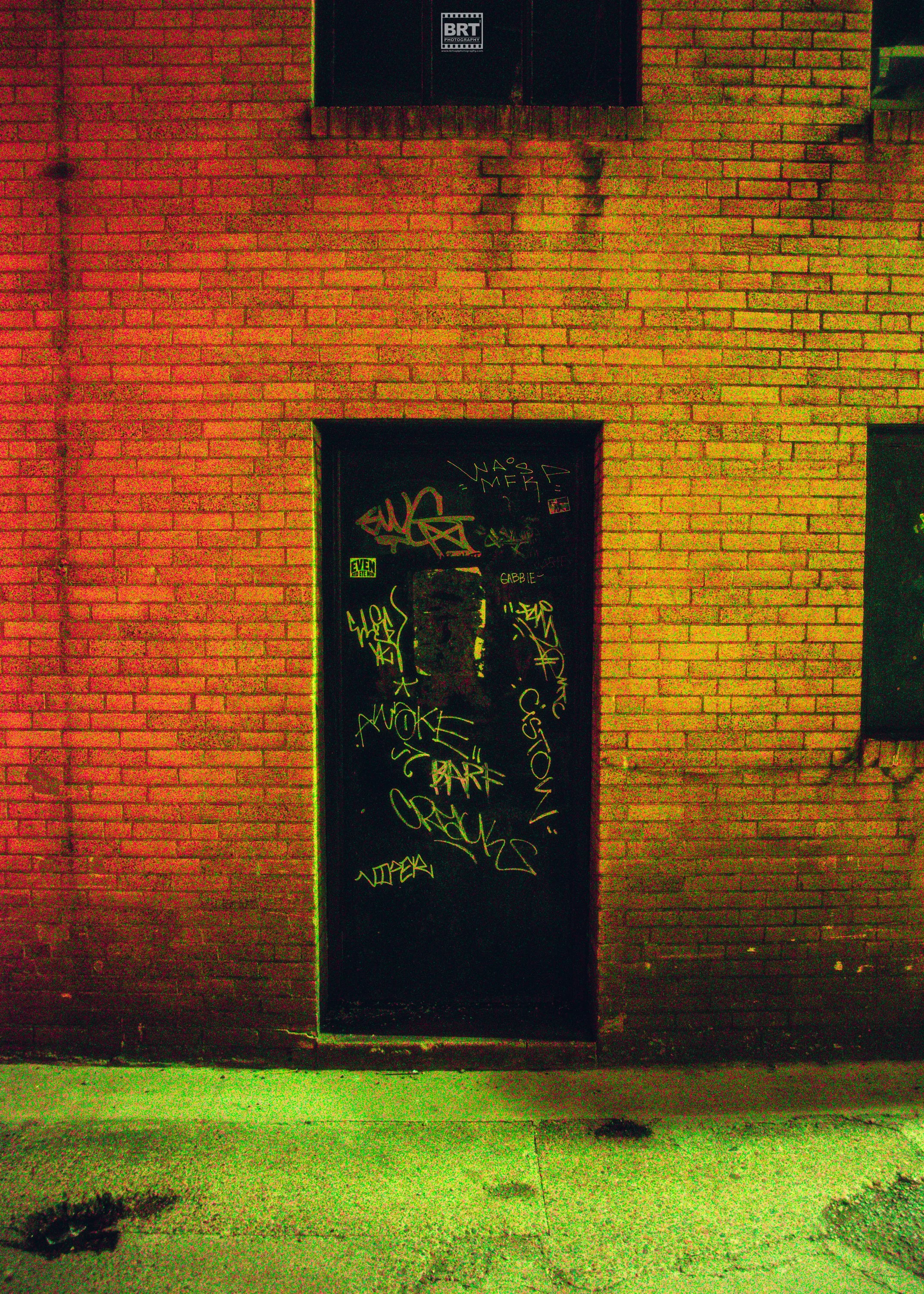 Black door covered in yellow graffiti tags and stickers in front of a brick wall.
