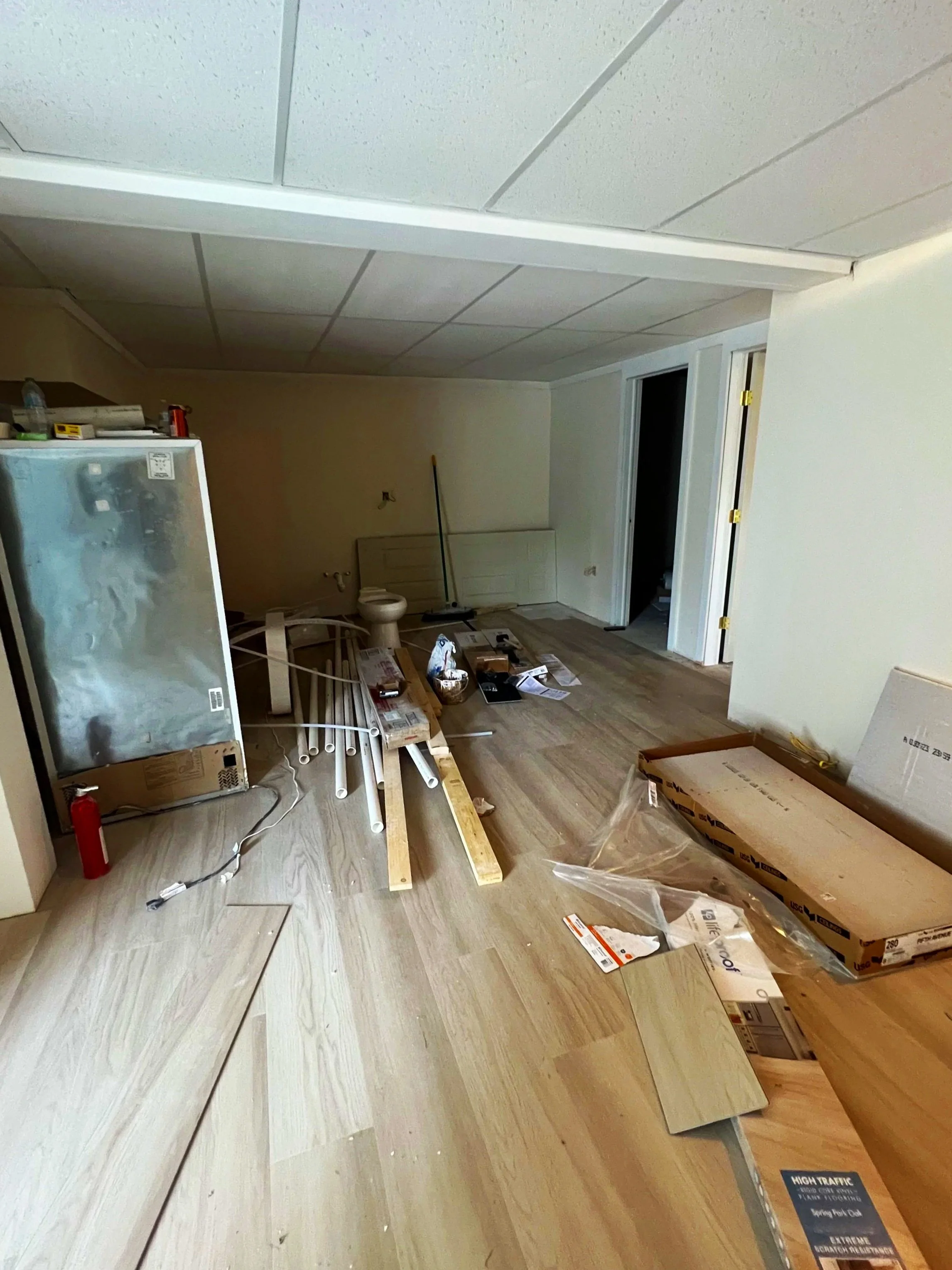 Room under renovation with scattered construction materials, including floor tiles, pipes, wood planks, and tools. A small refrigerator is visible on the left, and a doorway to another room is in the background.
