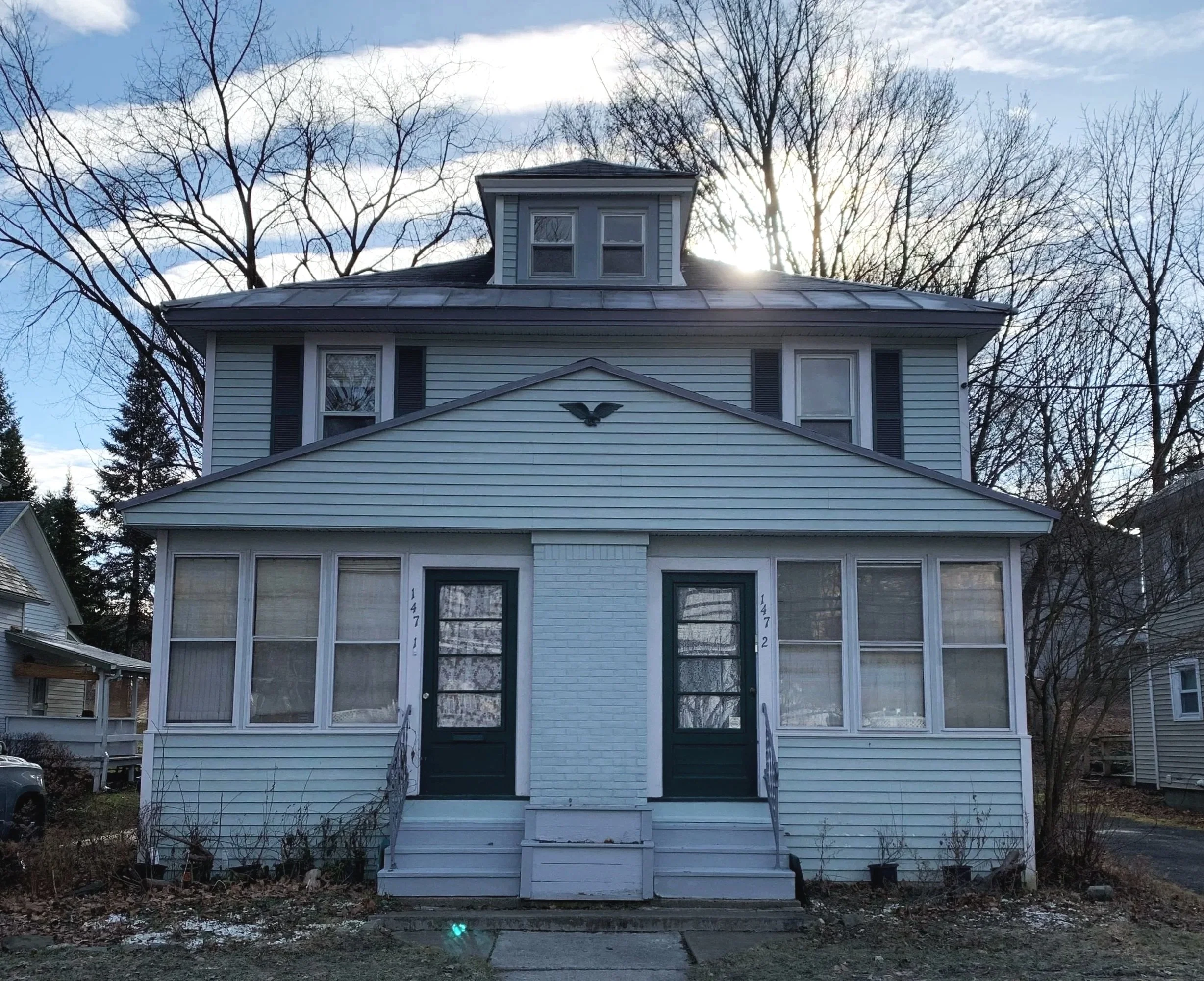 A three-story house with a small upper window dormer, white siding, dark green doors, and steps leading to the porch, with leafless trees and a blue sky with clouds and the sun shining behind the trees.