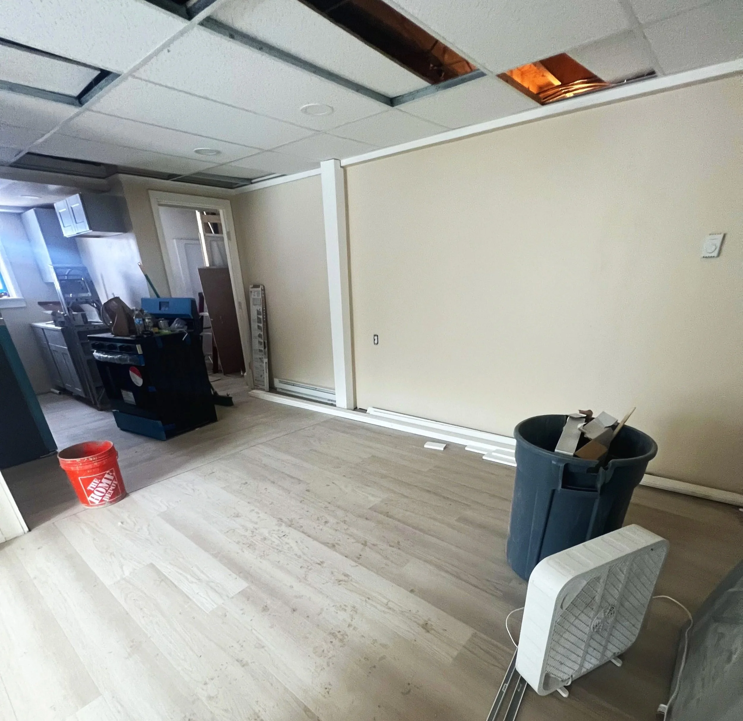 A partially renovated room with a dropped ceiling, some ceiling tiles removed, exposing wooden beams. The room has light-colored wooden flooring and a beige wall. There is a trash can, a space heater, and construction materials scattered around, indi