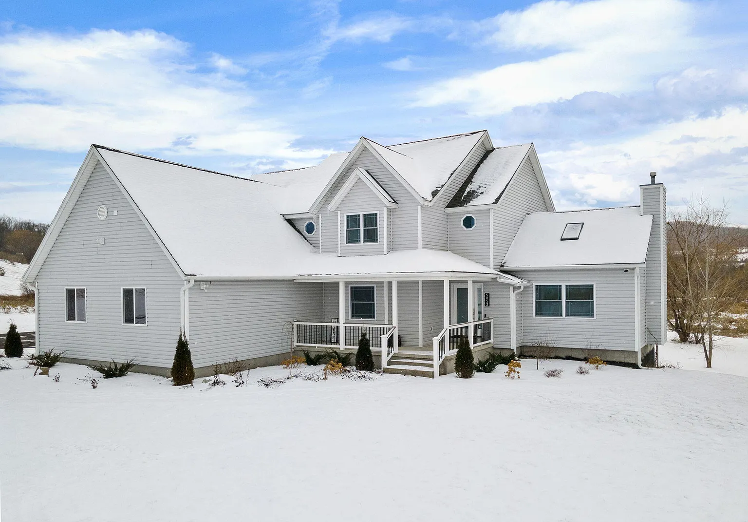 White house with multiple gabled roofs, covered porch, snowy yard, and leafless trees in background under cloudy blue sky.