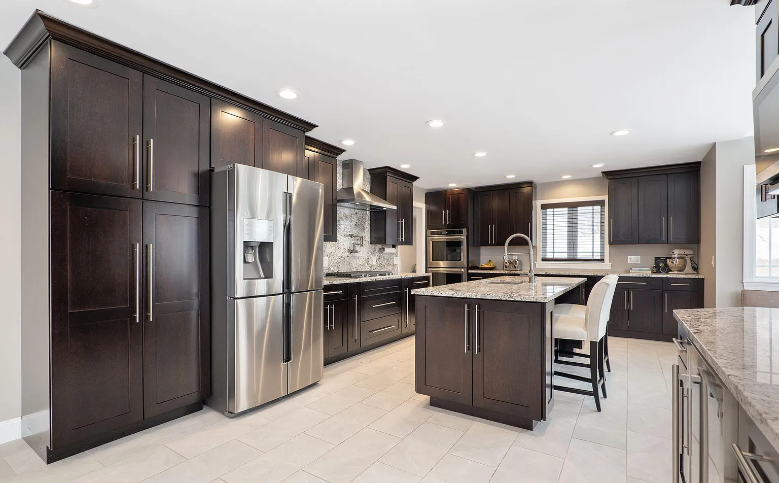 Modern kitchen with dark wood cabinets, stainless steel refrigerator, granite countertops, and tiled flooring.