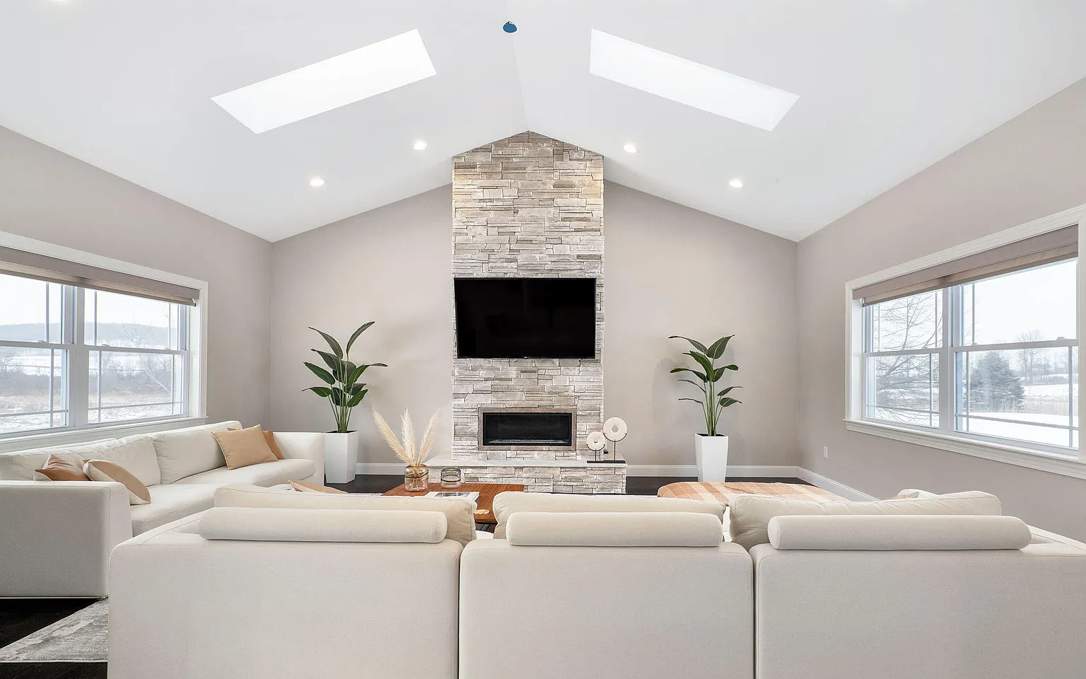 Living room with beige sofas, two large windows, a stone fireplace with a mounted TV, and indoor plants.
