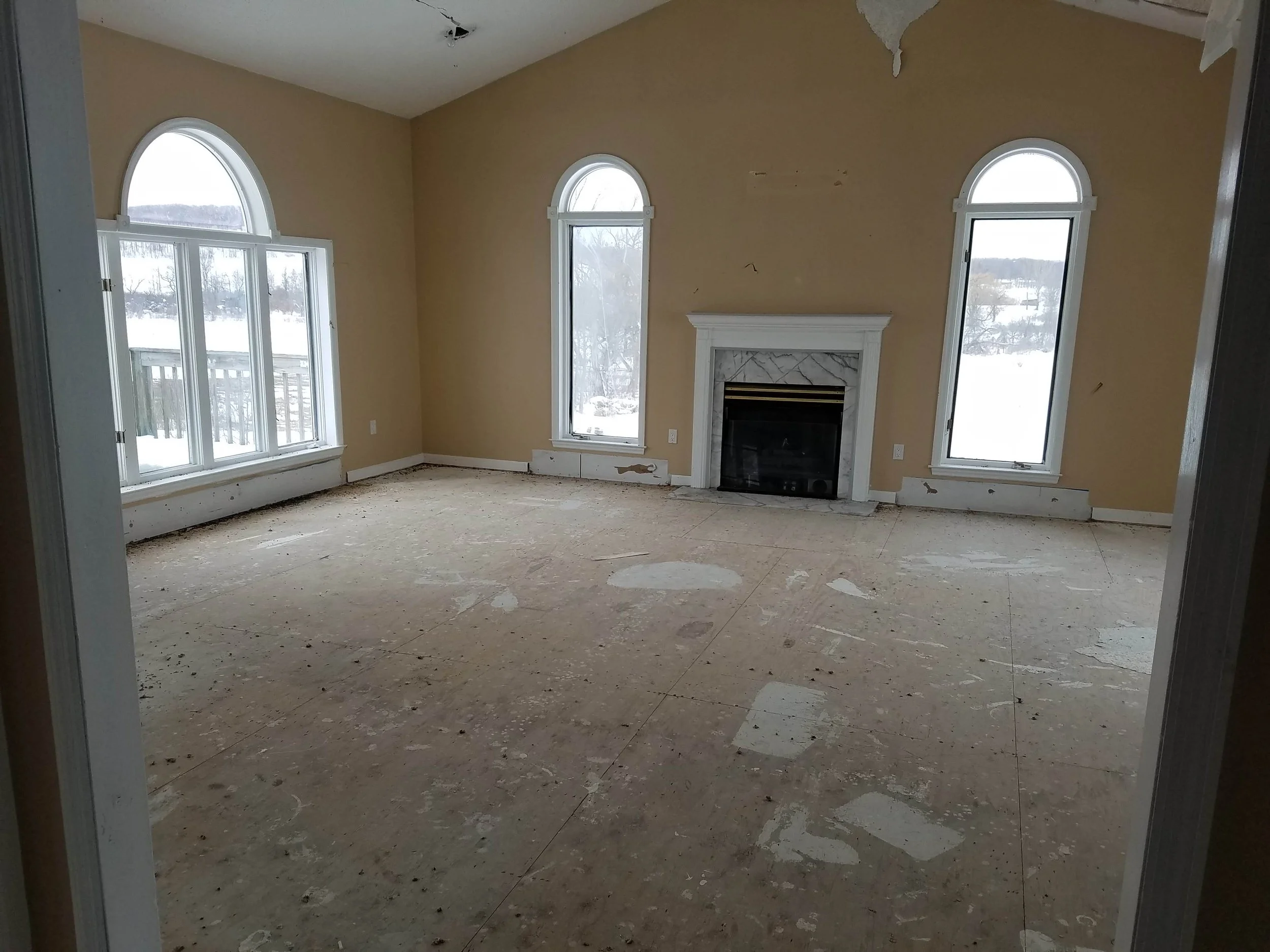 Empty living room with three large arched windows, a fireplace with marble surround, and unfinished flooring. Snow is visible outside through the windows.