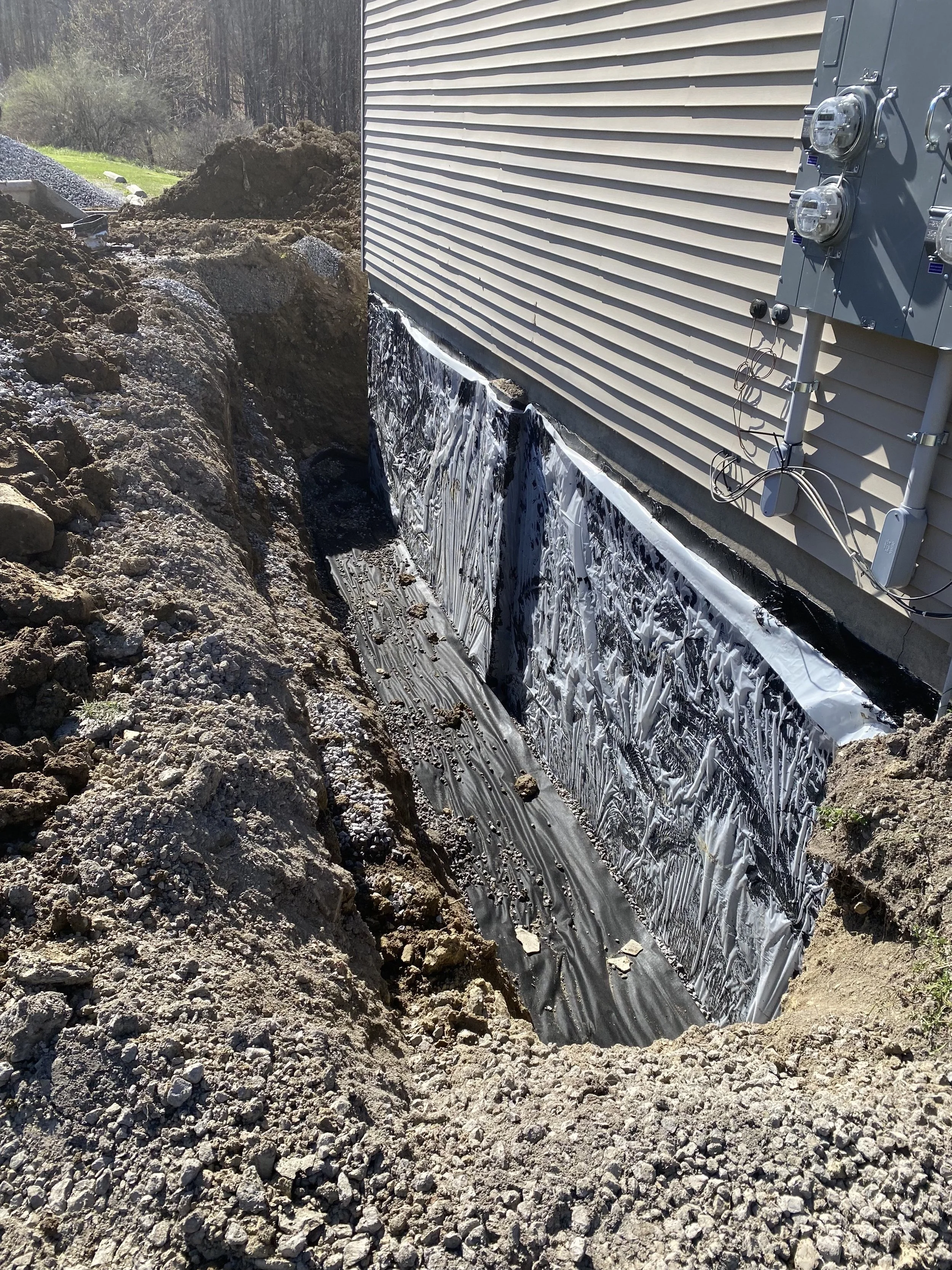 Excavated trench around the foundation of a house with black waterproofing membrane applied to the bottom and sides. Soil walls surround the trench, and an electrical meter box is mounted on the house's siding.