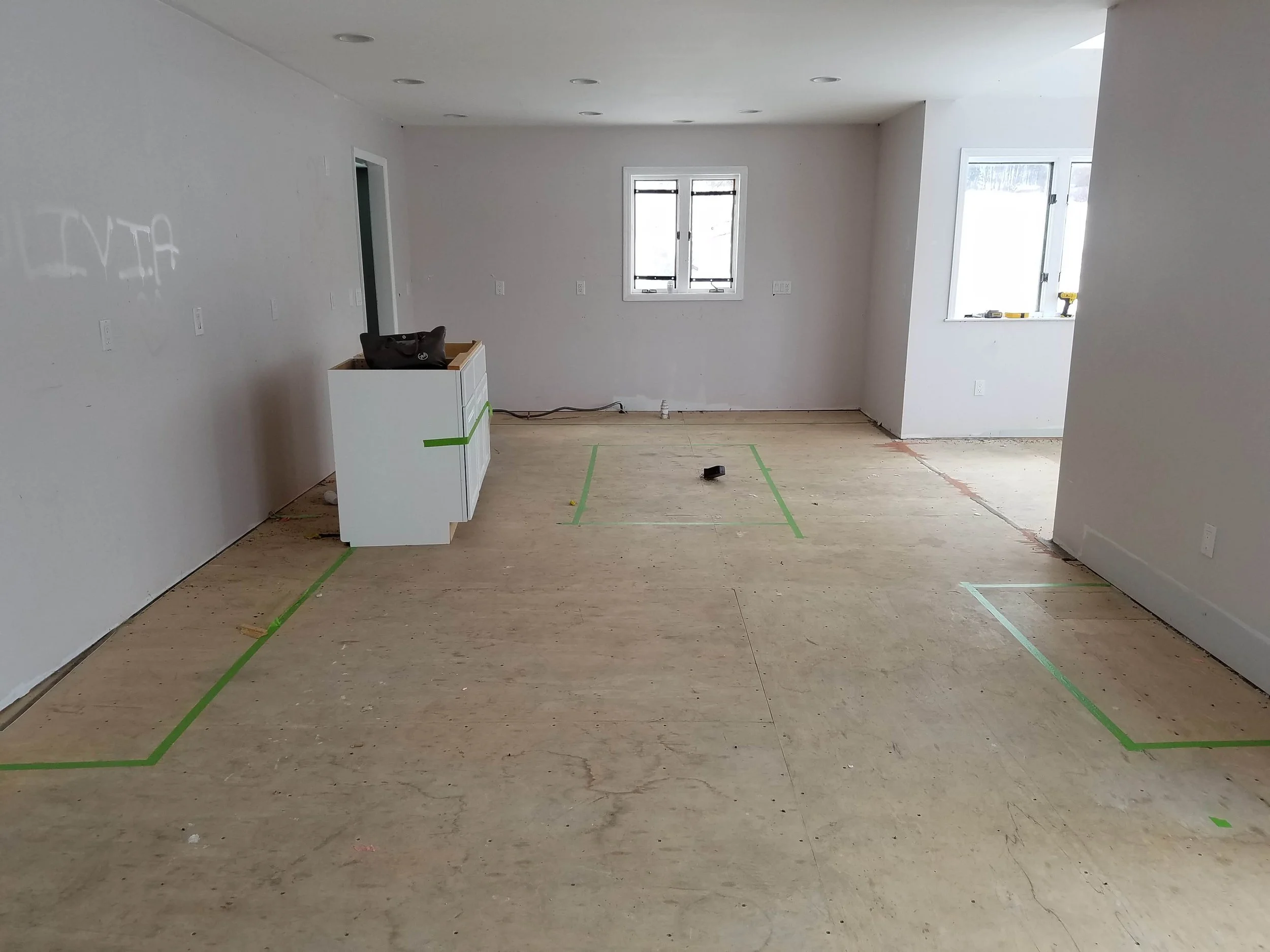 Empty room under construction with green painter's tape marking layout on the wooden floor, white walls, two windows, and a small white cabinet with black bag on top.