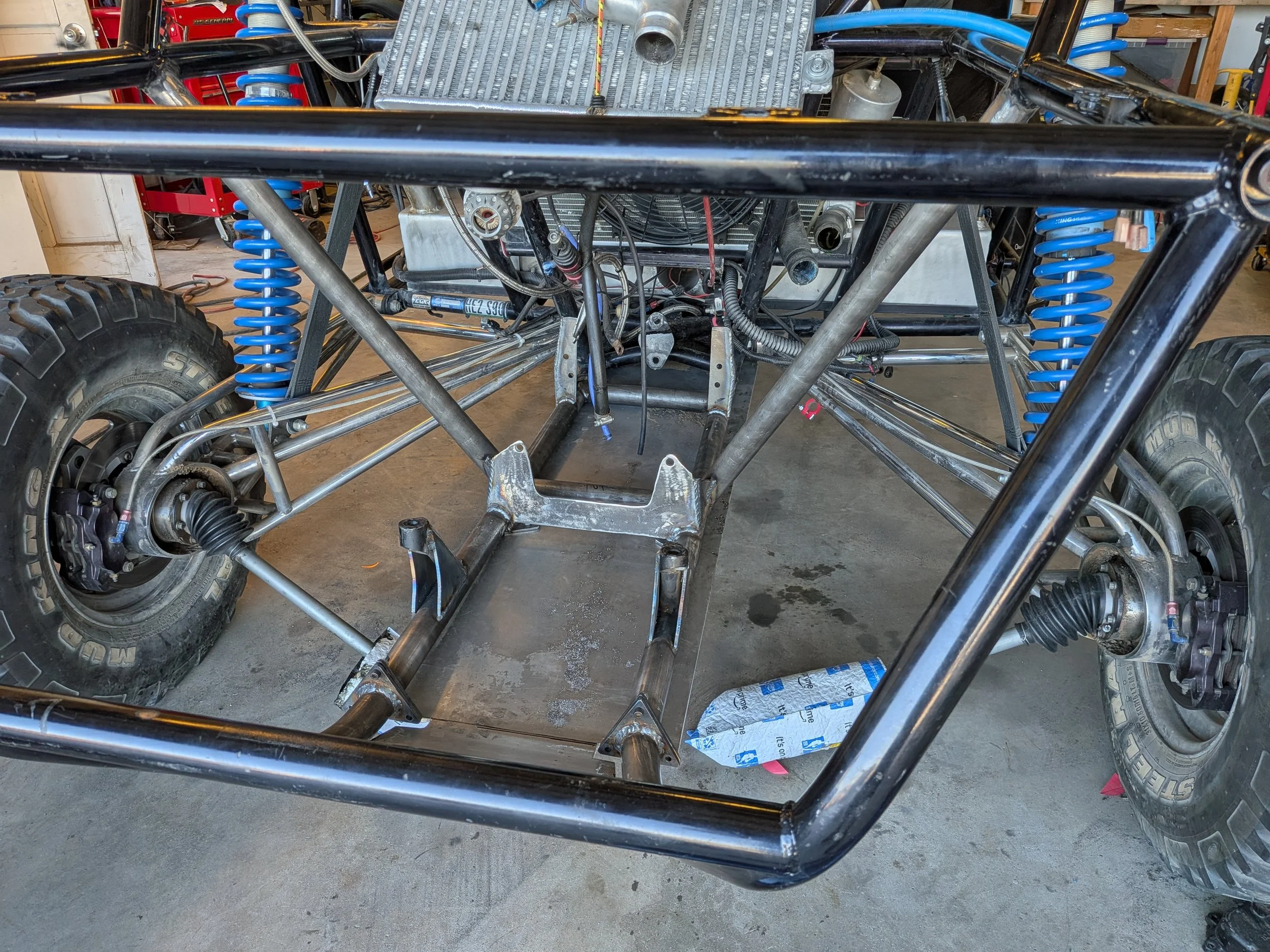 Partially assembled off-road racing vehicle with exposed chassis, blue coil shocks, and large off-road tires.