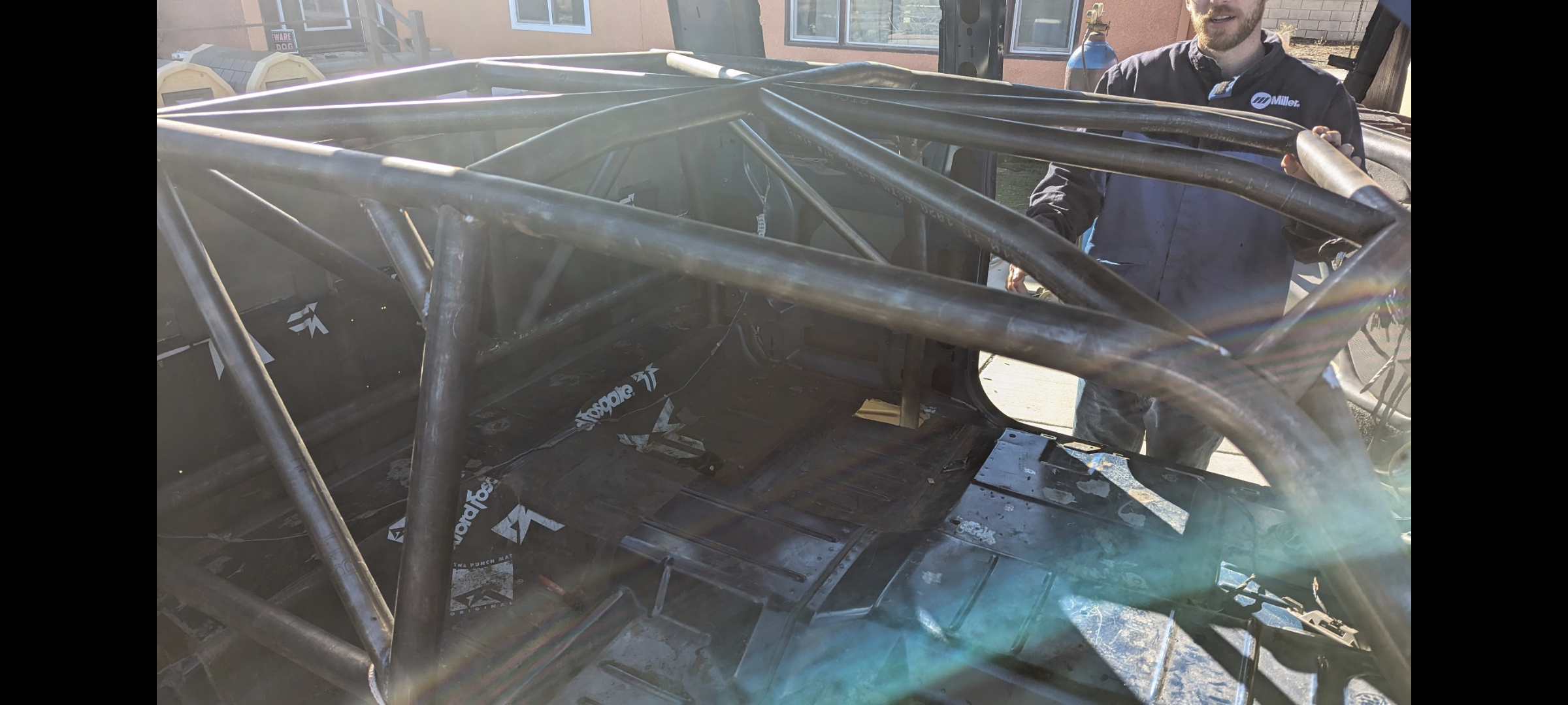 Behind the scenes view of a race car interior under construction with a man standing next to it, showing the metal roll cage and bare interior.