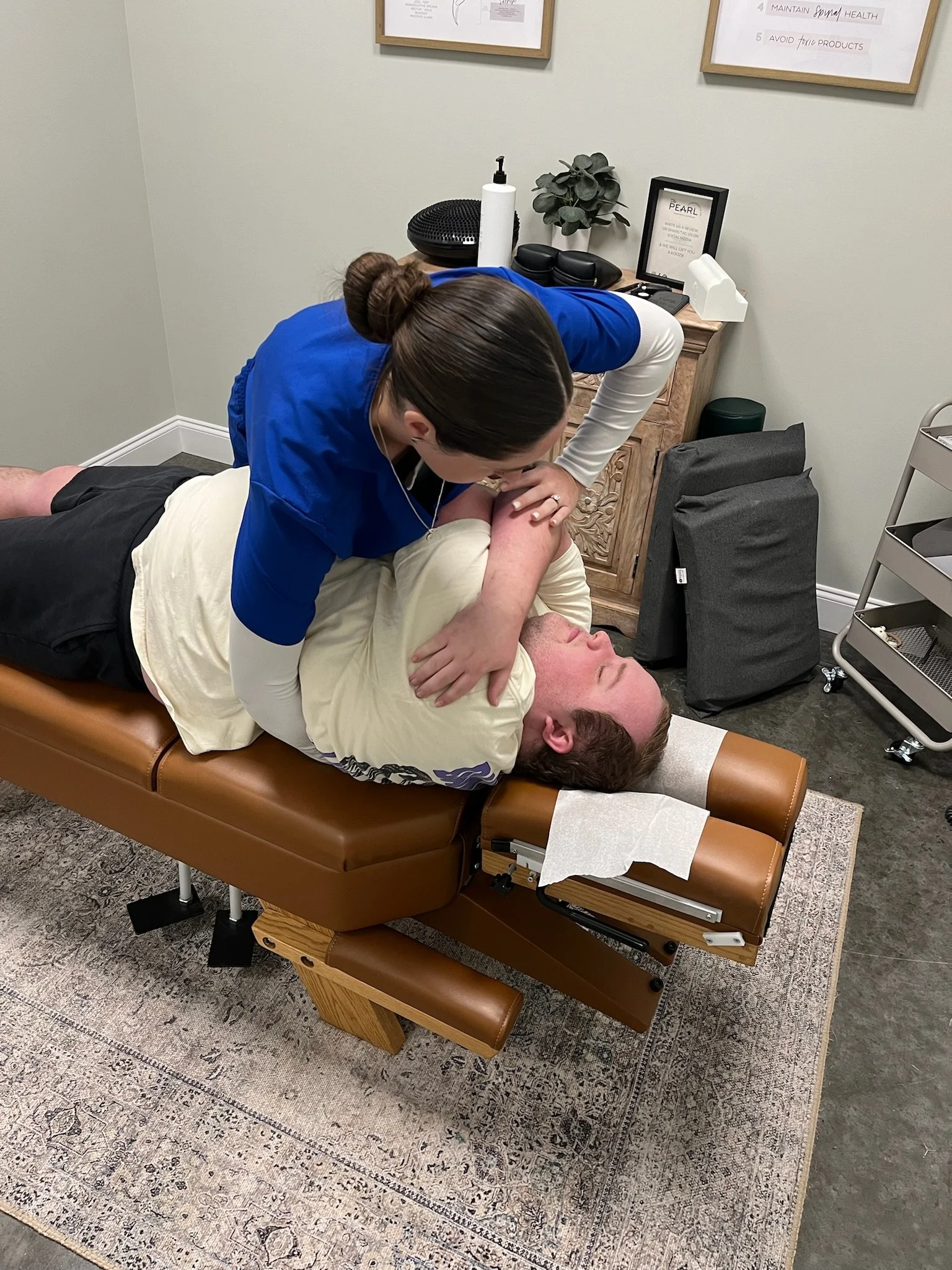 Medical professional performing an adjustment on a patient's shoulder in a clinic room with medical supplies and framed charts on the wall.