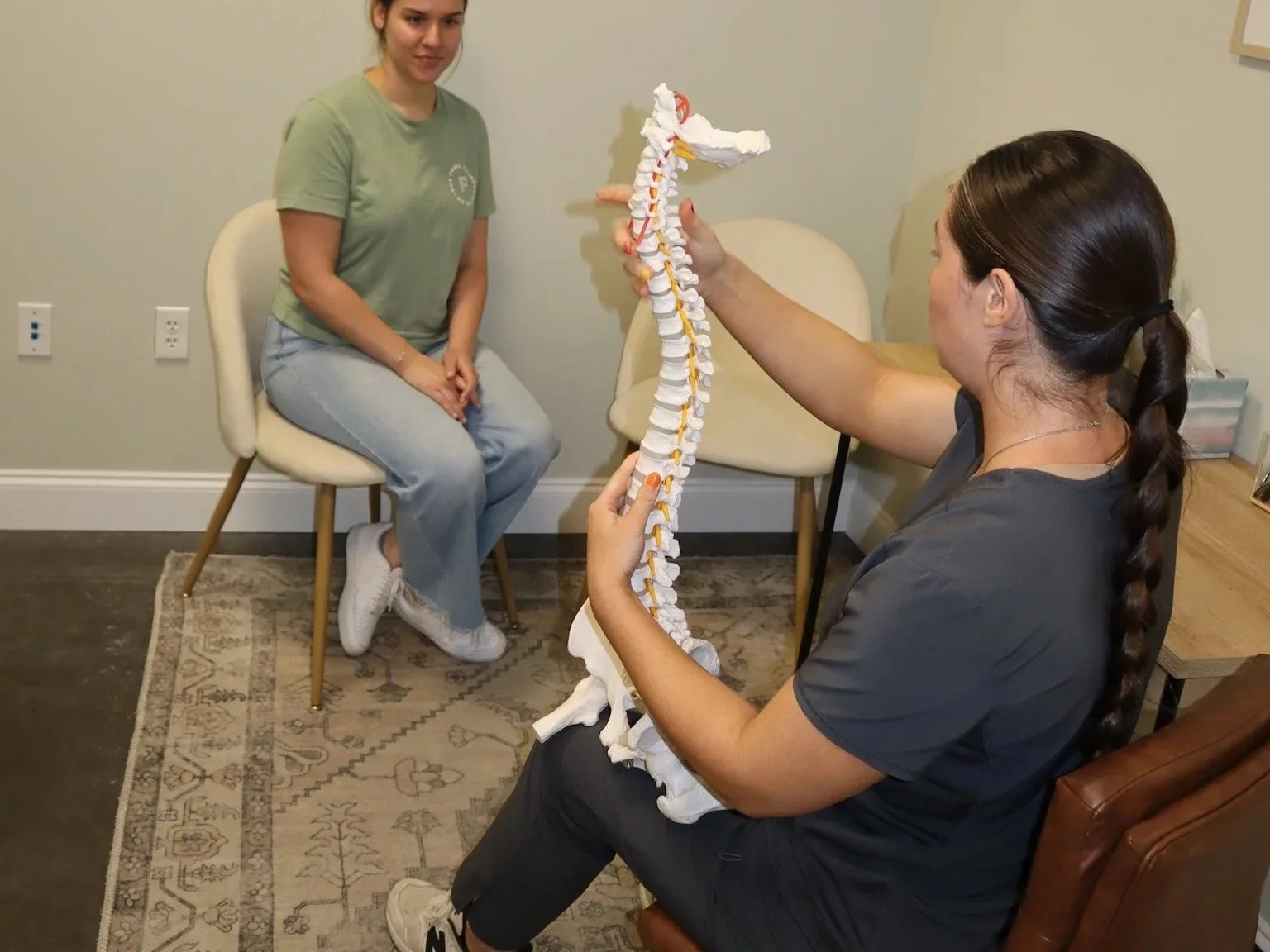Two women exam a model of a human spine in a medical consultation room. One woman is sitting on a chair, and the other is holding the spine model.