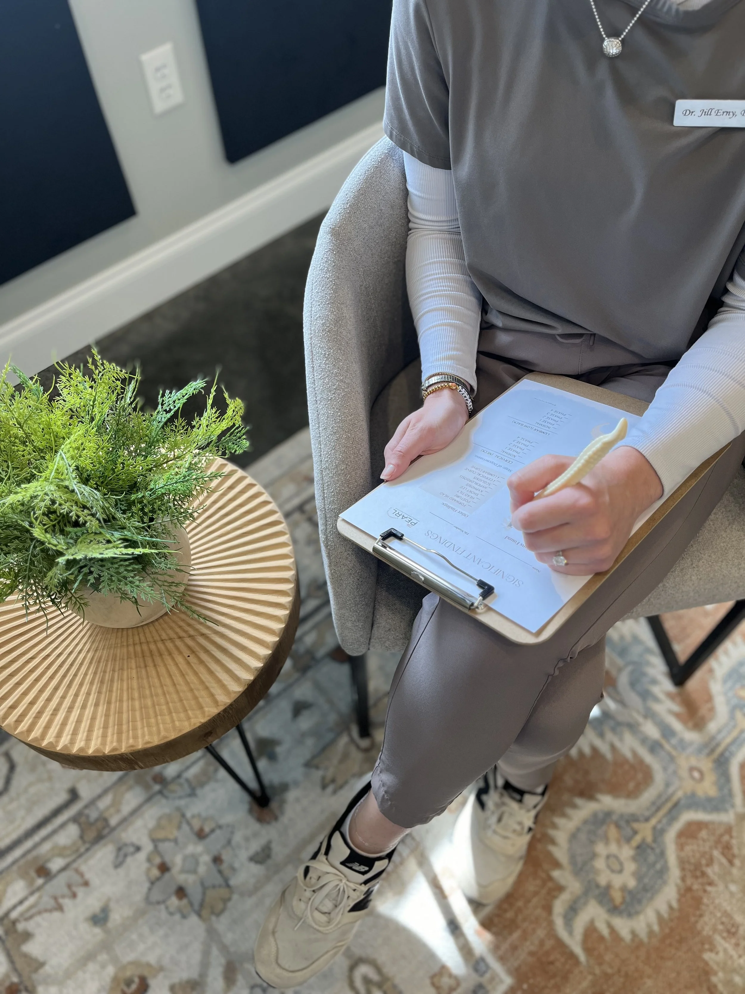 A person seated at a wooden table with a plant in a basket, writing on a clipboard with a pen. The person is wearing a gray medical uniform, a name badge, jewelry, and sneakers, with a patterned rug visible under the table.