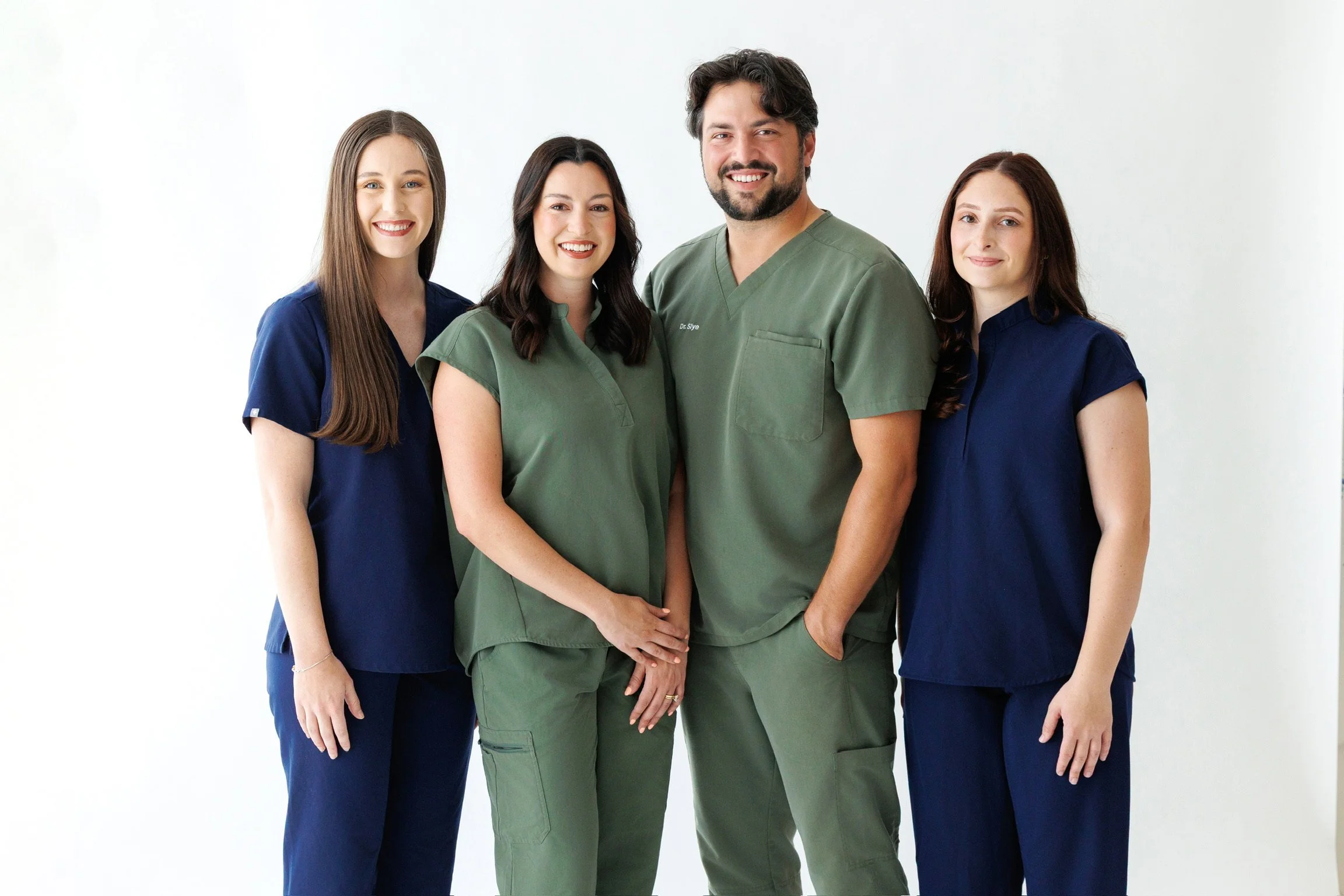 Group of four healthcare professionals, including a male doctor in green scrubs and three female nurses in dark blue scrubs, standing against a plain white background and smiling at the camera.
